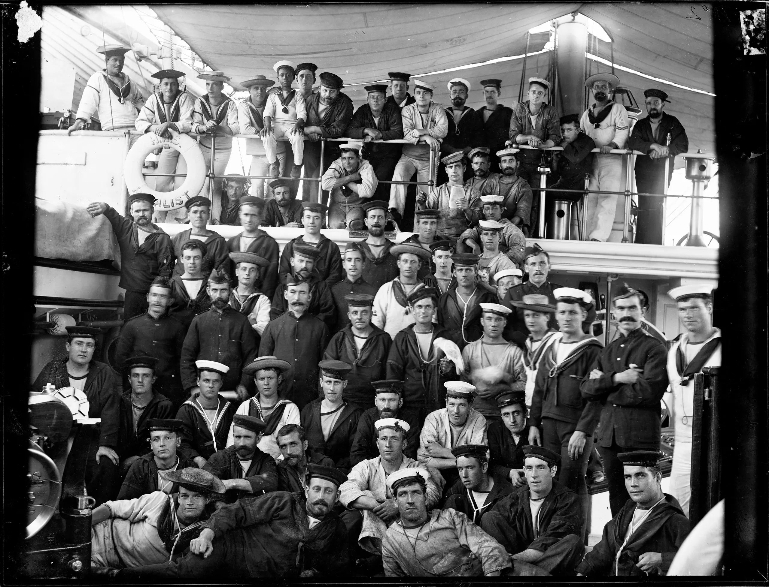 Glass plate negative, depicting ship's company of Royal Navy's Australia Station warship HMS Royalist. Landscape-format, black and white glass plate negative depicting a large group of sailors in uniform, all wearing caps or hats, on two decks of a naval ship. The ship is HMS Royalist, Imperial Squadron, Australia Station.