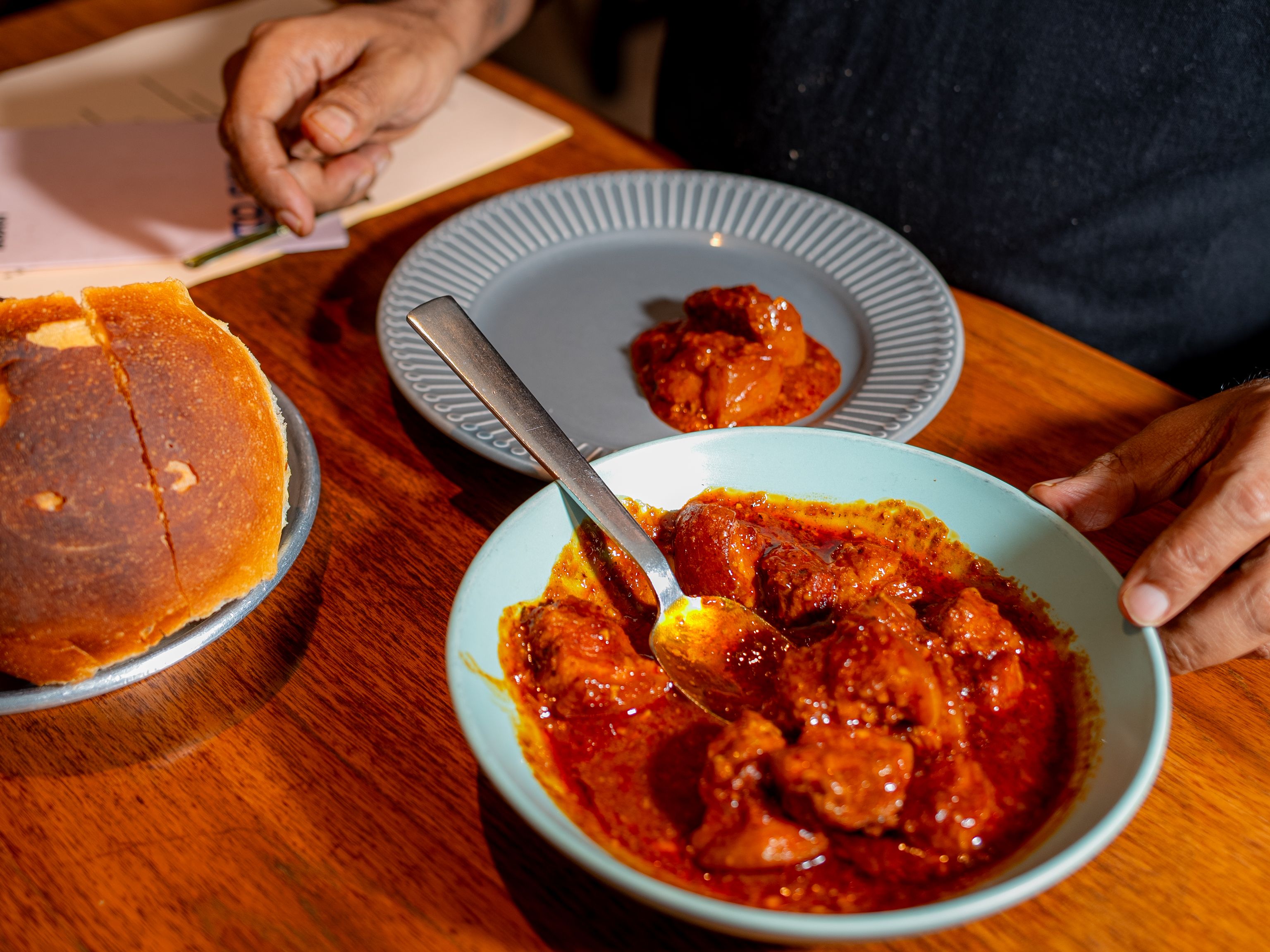A plate of bread, a plate with a spoonful of red-coloured curry and a bowl of the same curry on a polished wood tabletop.
