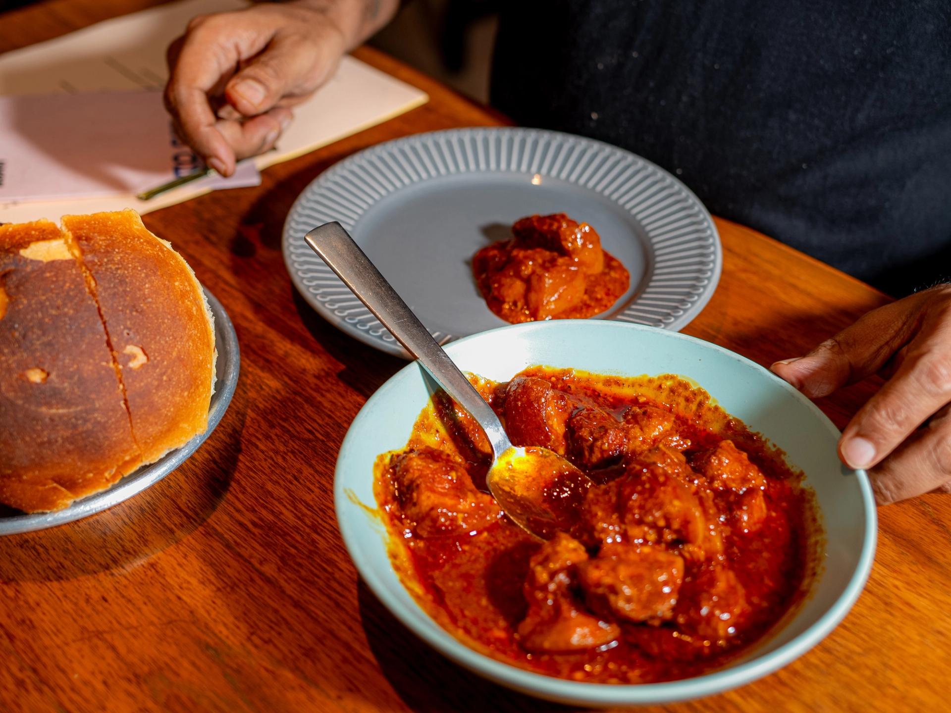 A plate of bread, a plate with a spoonful of red-coloured curry and a bowl of the same curry on a polished wood tabletop.