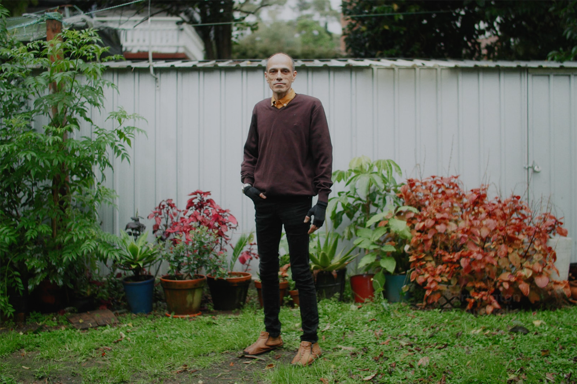 A figure stands in suburban garden wearing a maroon jumper and black jeans.