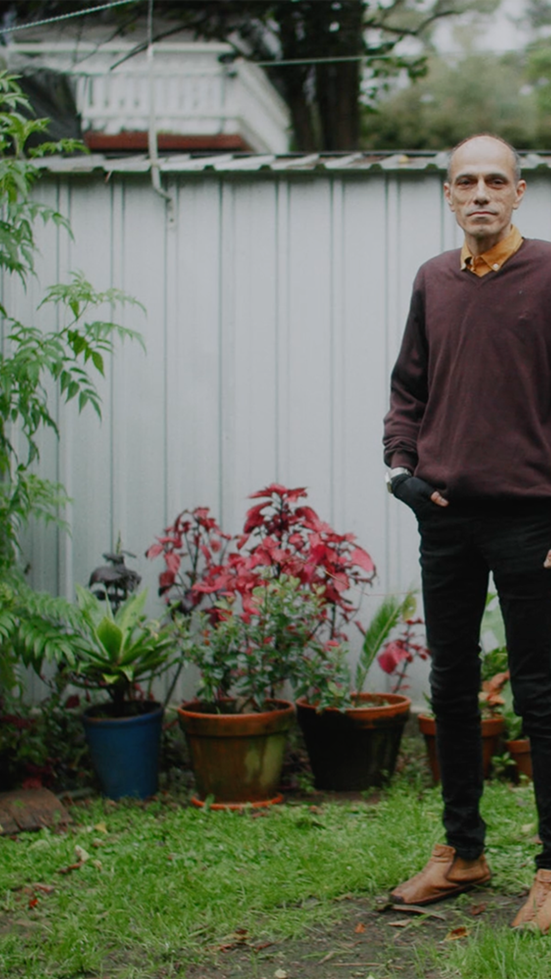 A figure stands in suburban garden wearing a maroon jumper and black jeans.