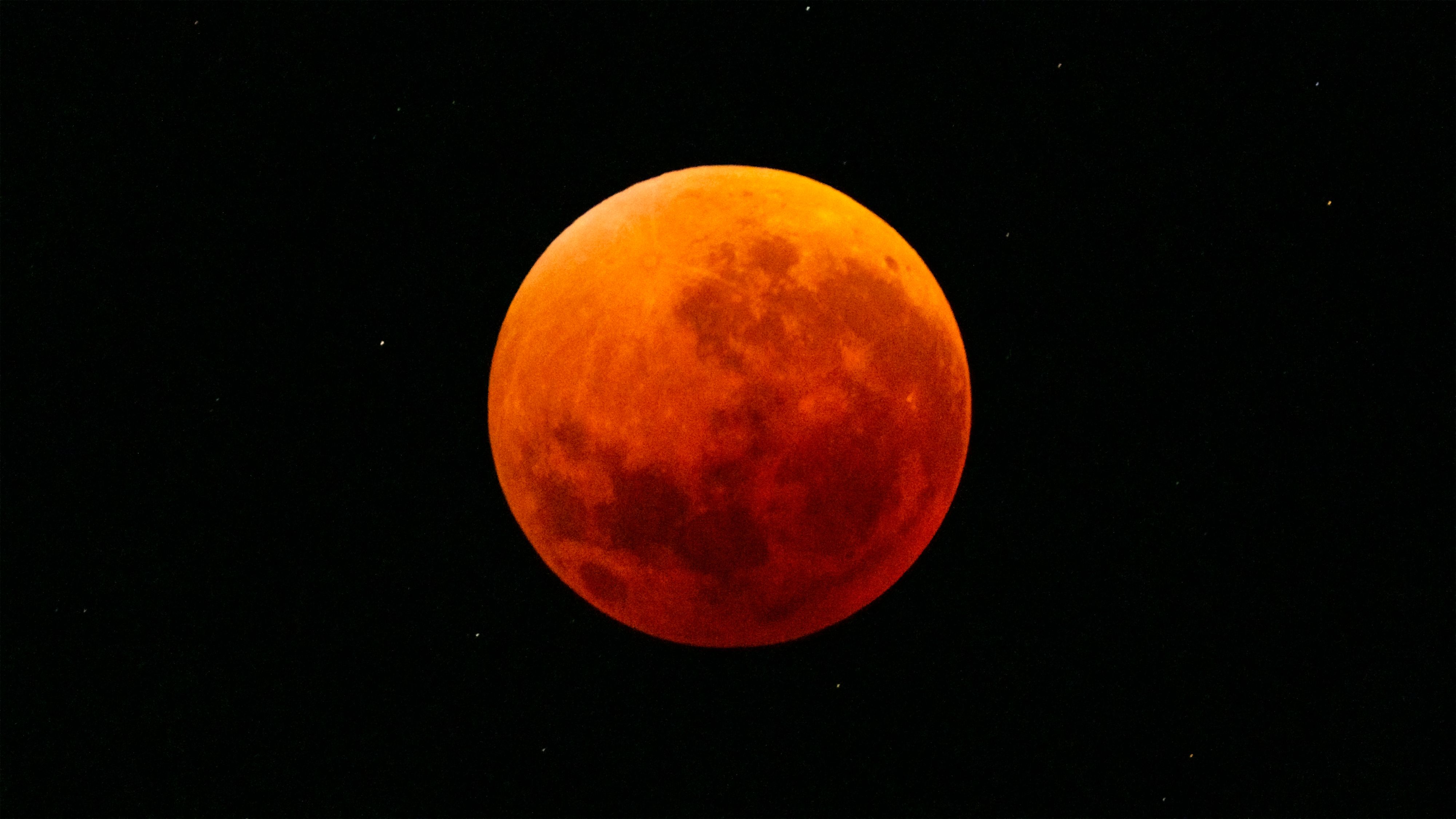 Total lunar eclipse showing the Moon fully immersed in Earth's shadow, appearing deep orange-red (blood moon) with visible surface details and a starry black background.
