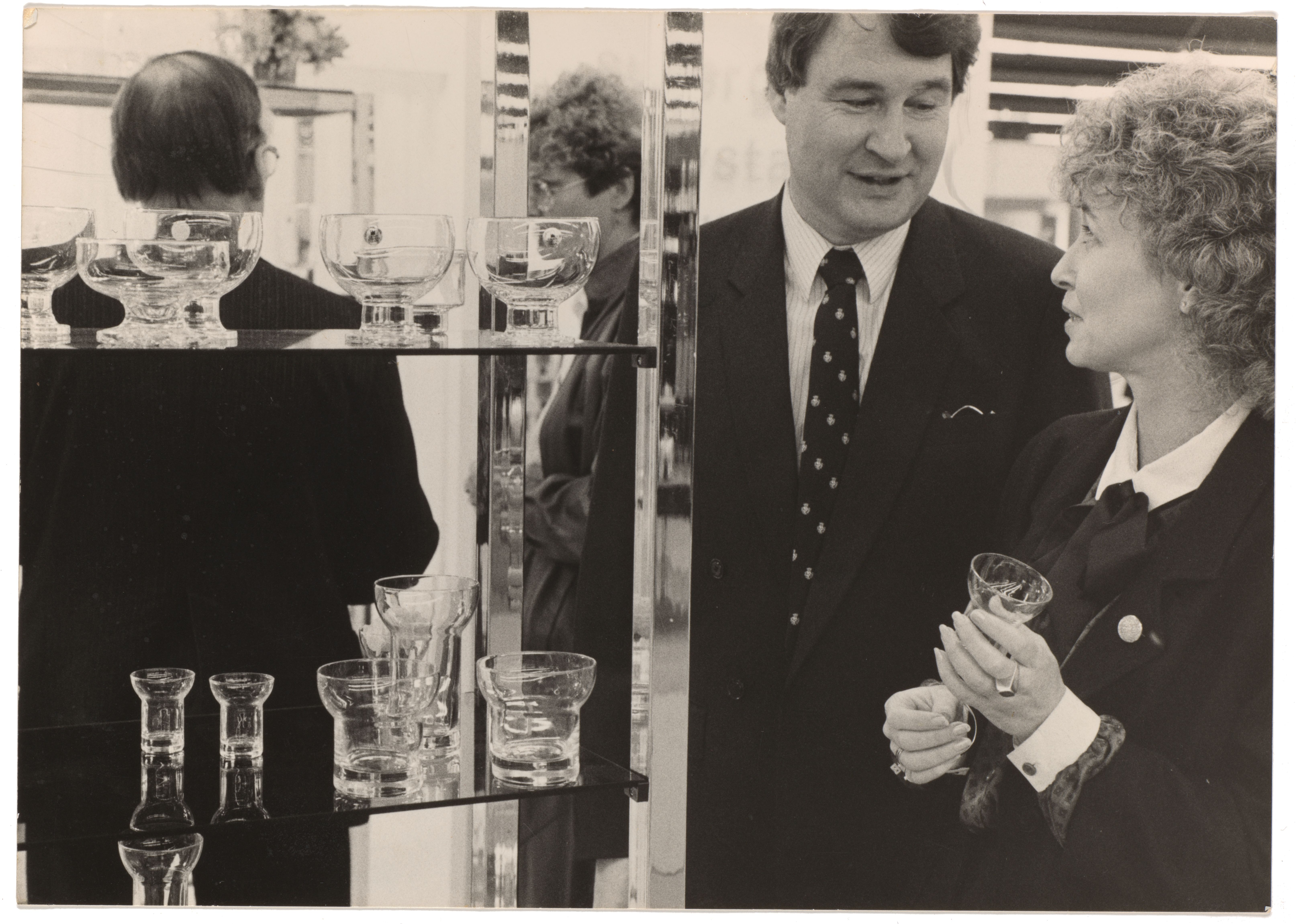 Black and white photograph of multiple glasses on a shelf, to the right designer Denise Larcombe and guest hold one of the glasses at the Frankfurt Fair.