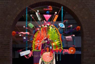 View of a mannequin from the waist up wearing a bold, colourful costume with large arch head piece. The headpiece has feathers, streamers, and other found materials sticking out from the main structure.