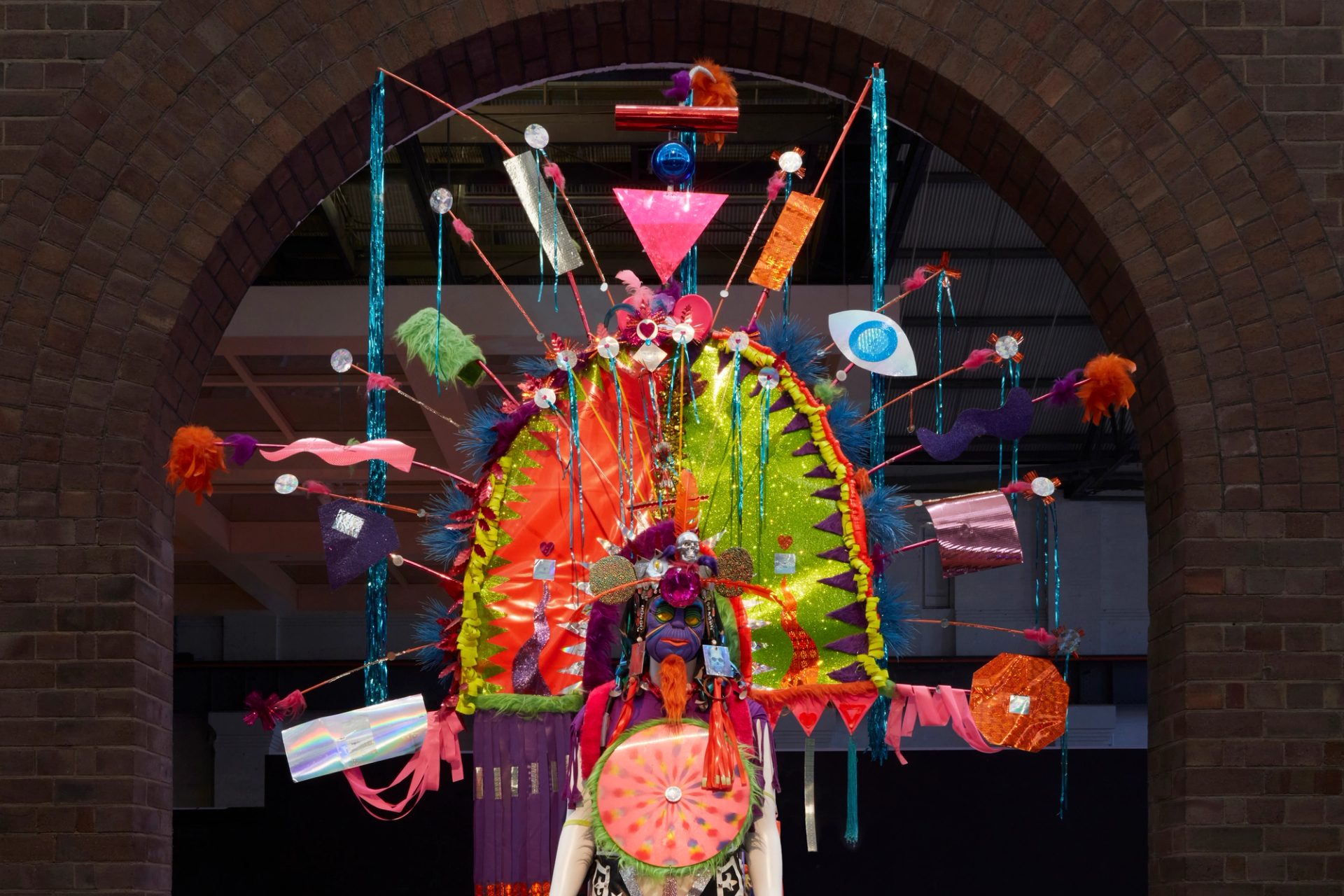 View of a mannequin from the waist up wearing a bold, colourful costume with large arch head piece. The headpiece has feathers, streamers, and other found materials sticking out from the main structure.