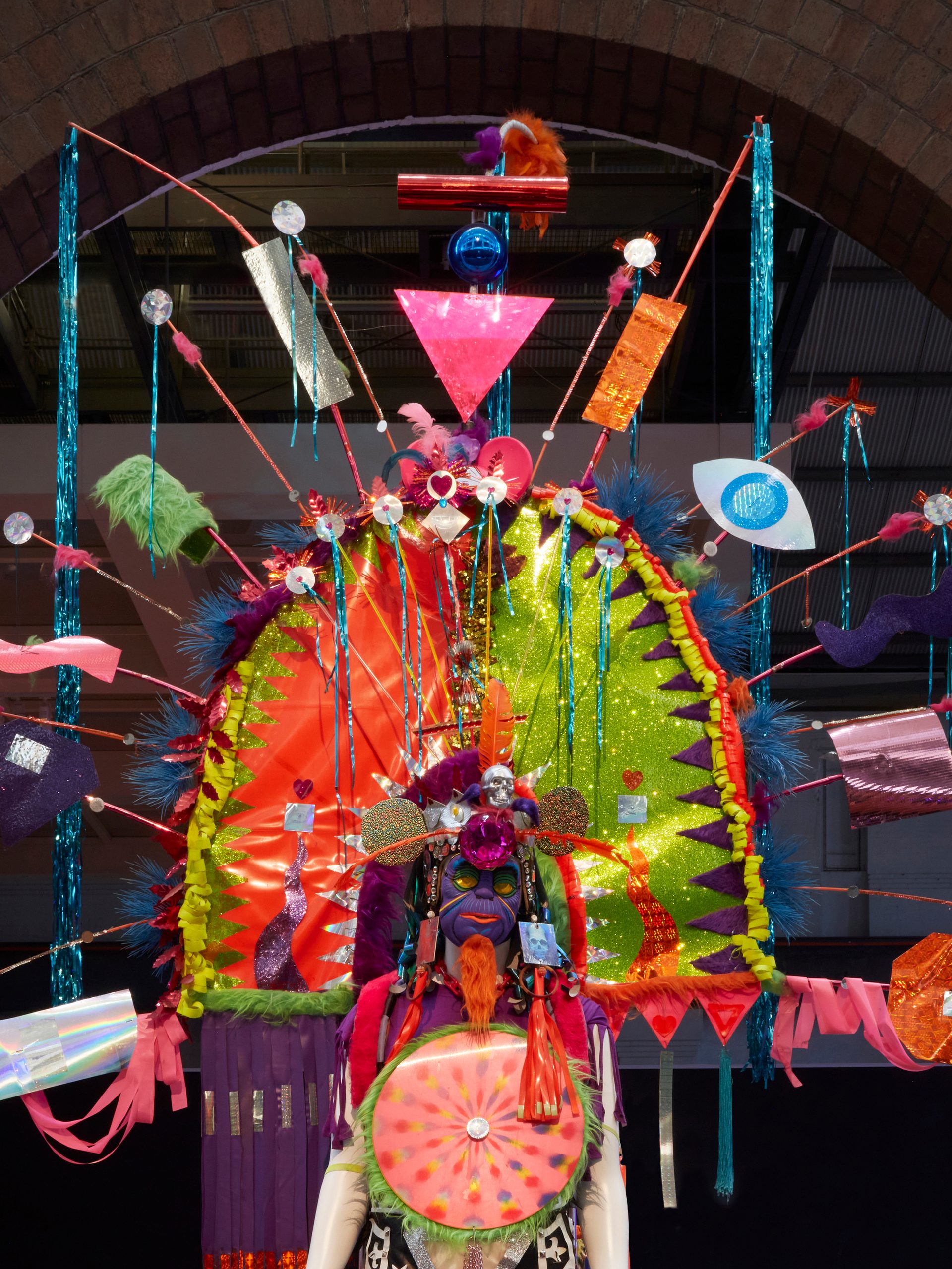 View of a mannequin from the waist up wearing a bold, colourful costume with large arch head piece. The headpiece has feathers, streamers, and other found materials sticking out from the main structure.