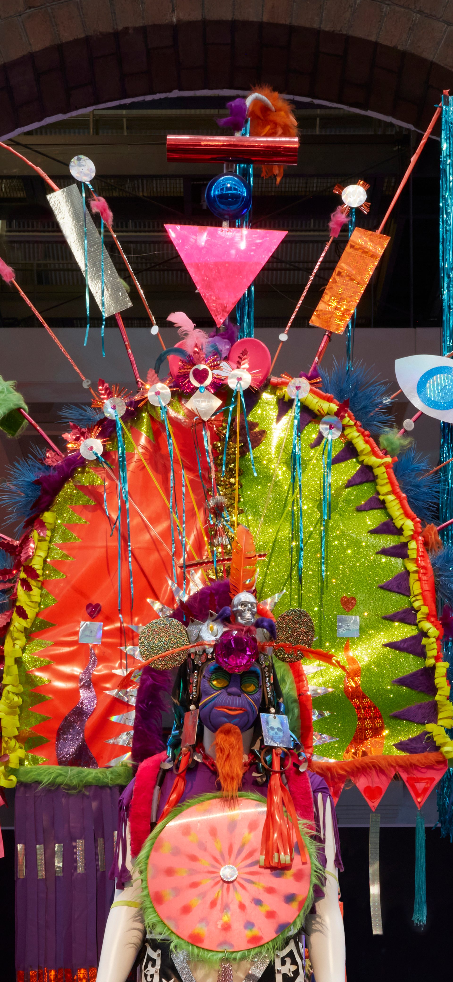 View of a mannequin from the waist up wearing a bold, colourful costume with large arch head piece. The headpiece has feathers, streamers, and other found materials sticking out from the main structure.