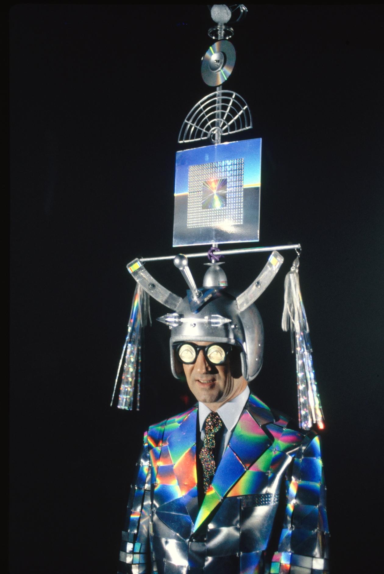 A figure wearing a large silver headpiece, black rimmed glasses and a navy suit with a rainbow gradient pattern.