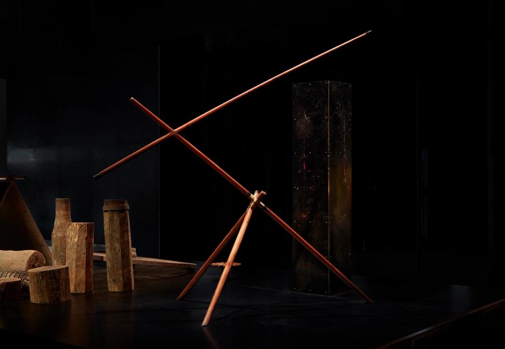 A series of interconnected dark wood beams. The sculpture is against a dark black background and is spotlit.