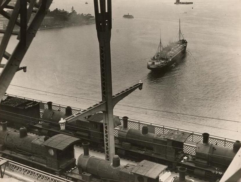 Black and white photo of five small black locomotives on a section of Sydney Harbour Bridge with cargo ship on the water below.
