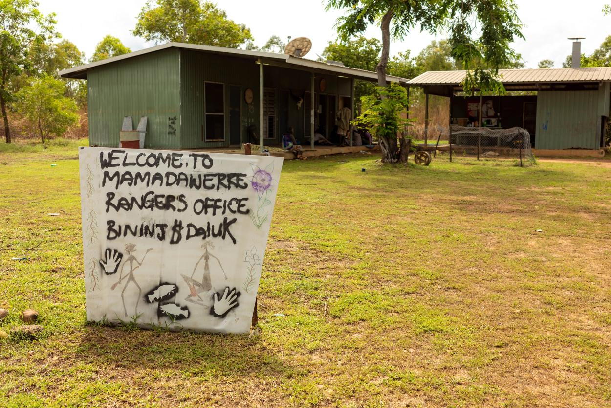 A white sign with black text reading, ‘WELCOME TO MAMADAWERRE RANGERS OFFICE BININJ$DAIUK,’ sits on short green grass in front of two small green tin offices.