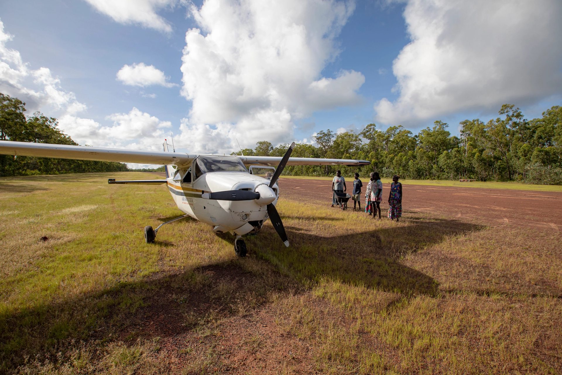 A small passenger plane in a large open field. There is a group of five people, one holding a wheelbarrow, to the plane’s left.