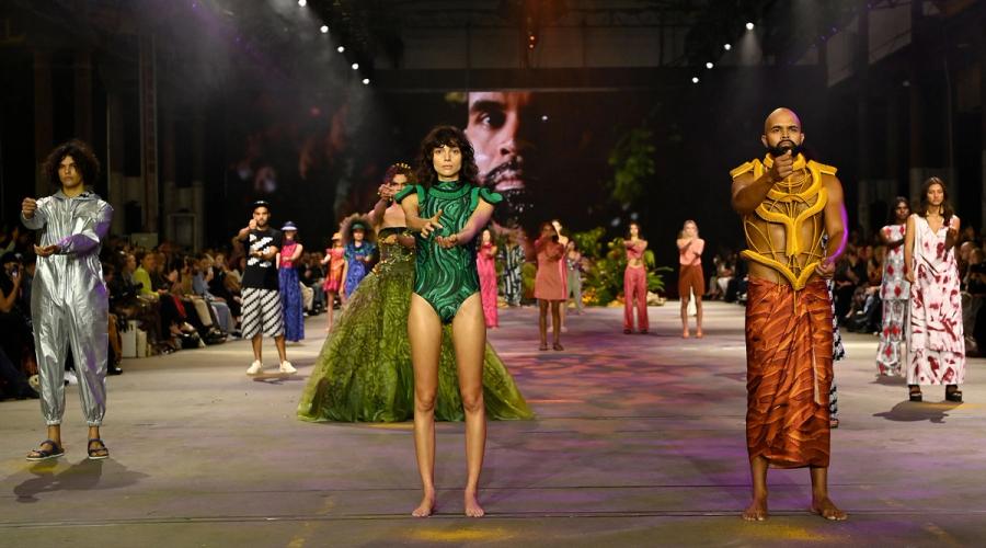 A wide fashion runway with Indigenous models dressed in various styles and colours of clothing and swimwear. At the front are a man and a woman. They are holding sand in one hand and letting it fall into the other hand.