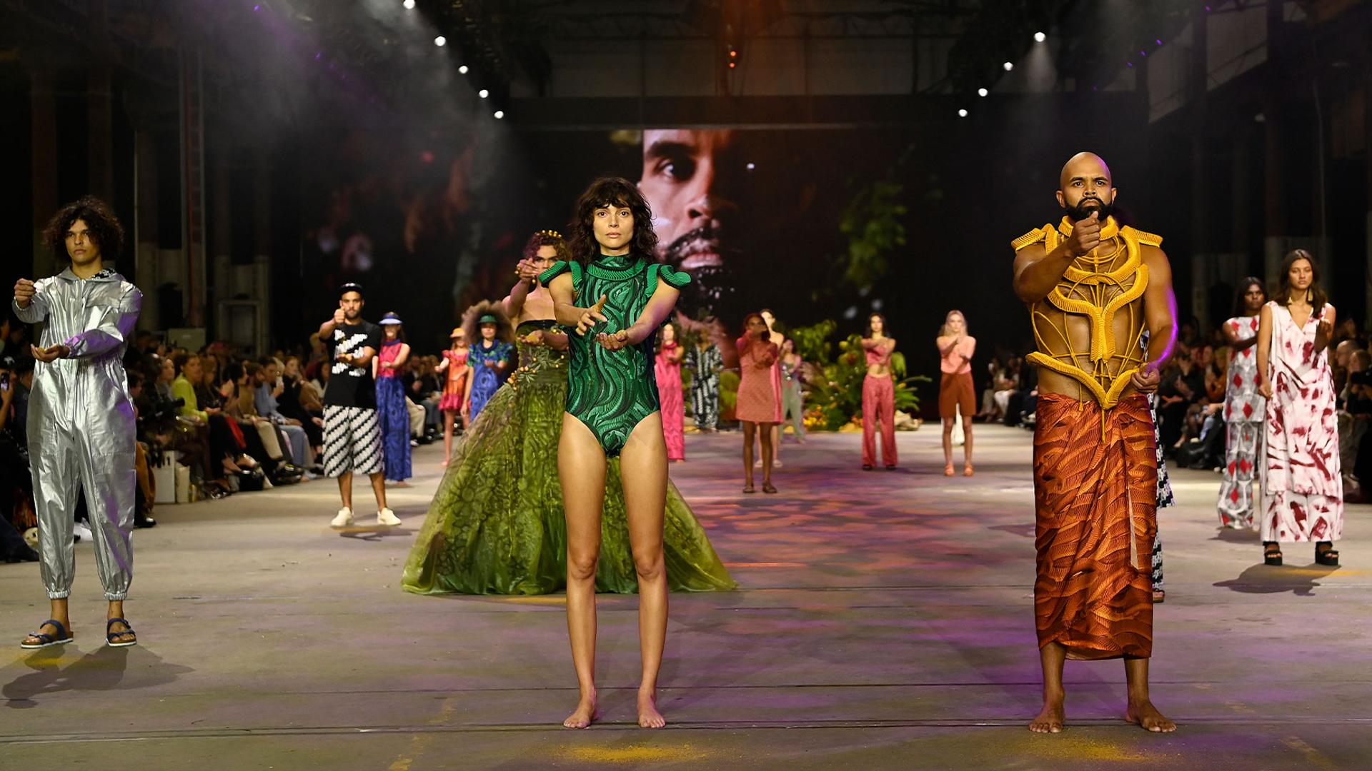 A wide fashion runway with Indigenous models dressed in various styles and colours of clothing and swimwear. At the front are a man and a woman. They are holding sand in one hand and letting it fall into the other hand.