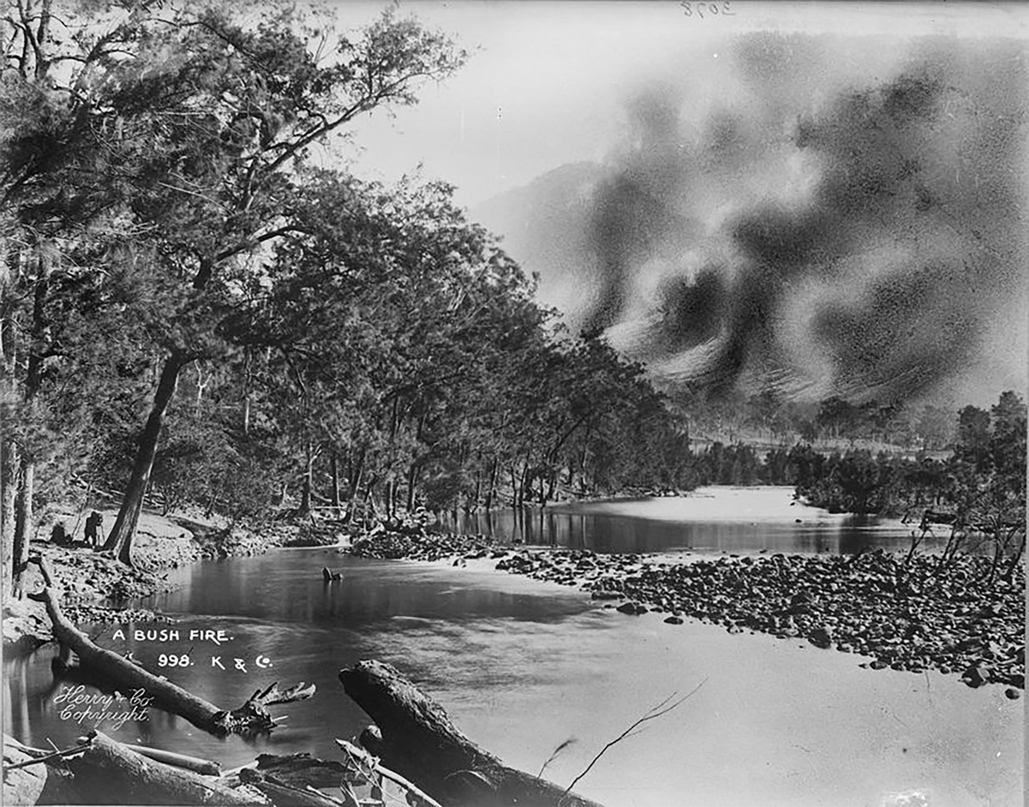 Black and white photographic print showing a river and a bushfire scene behind.