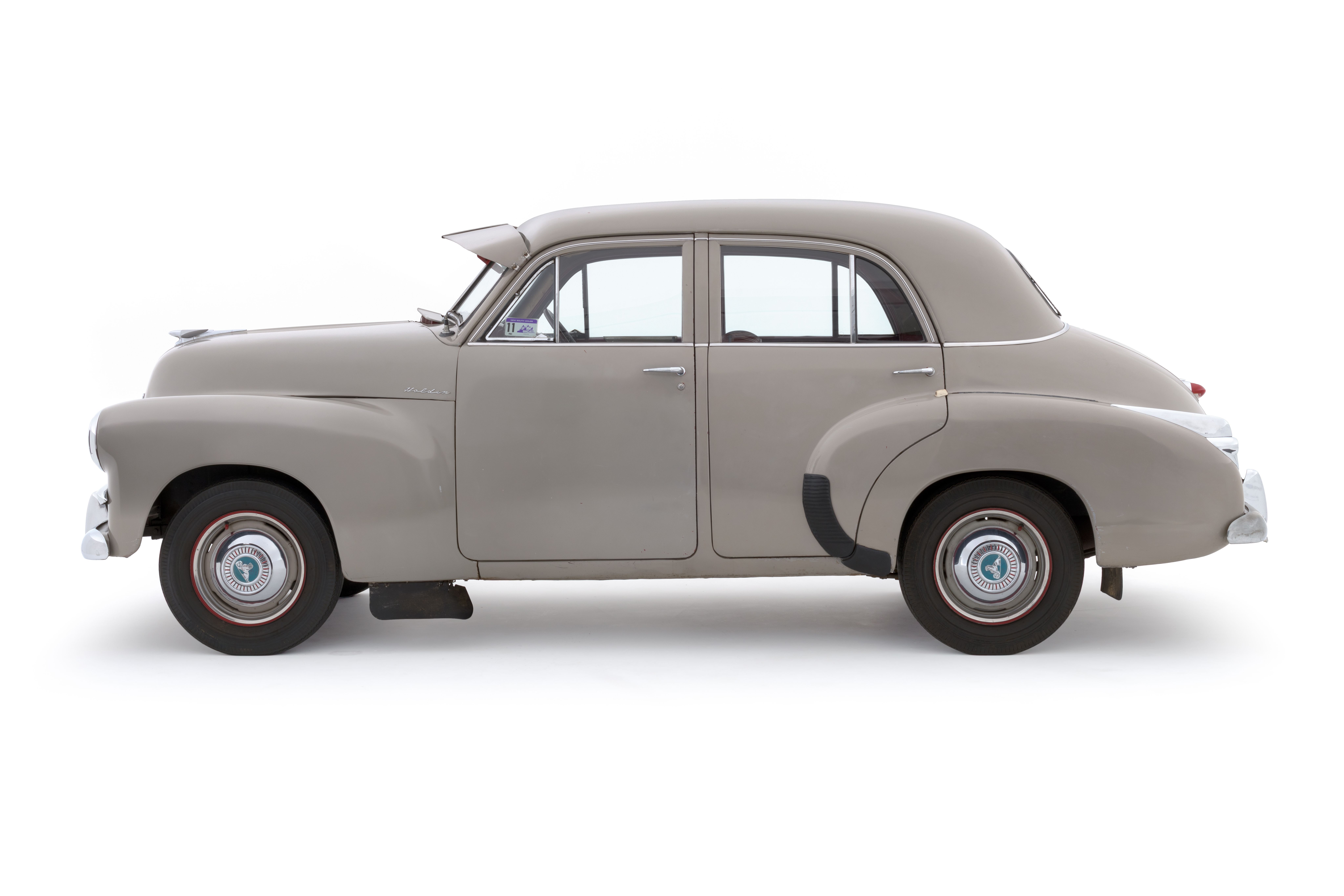 A light grey, beige car. A 1955 FJ225 Holden Special four door sedan featuring a pale grey exterior with chrome brightwork. The interior of the car comprises red leather upholstery for the seats, two-toned red and pale grey for the inner door trim, a red painted dashboard and red carpeted floor at the back.