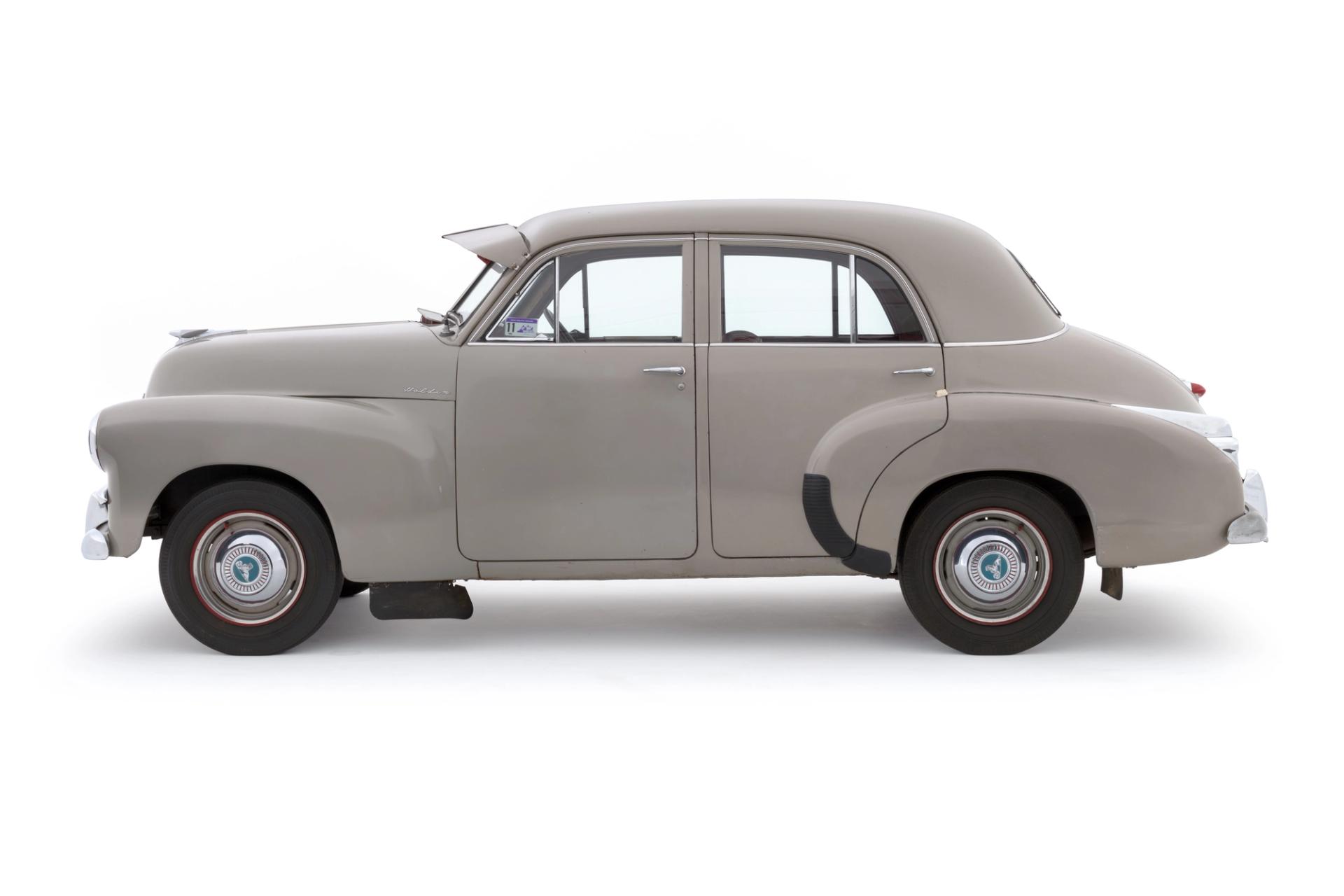 A light grey, beige car. A 1955 FJ225 Holden Special four door sedan featuring a pale grey exterior with chrome brightwork. The interior of the car comprises red leather upholstery for the seats, two-toned red and pale grey for the inner door trim, a red painted dashboard and red carpeted floor at the back.