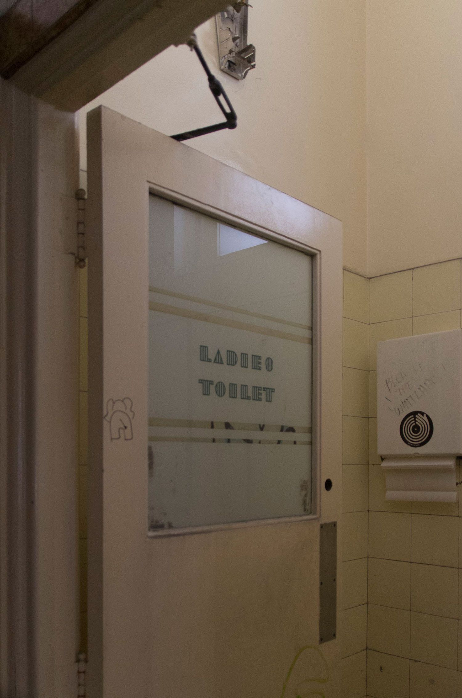 The bathroom door of a ladies bathroom in the old Clare Hotel. The surroundings are dirty and old. The text on the door reads ‘LADIES’.