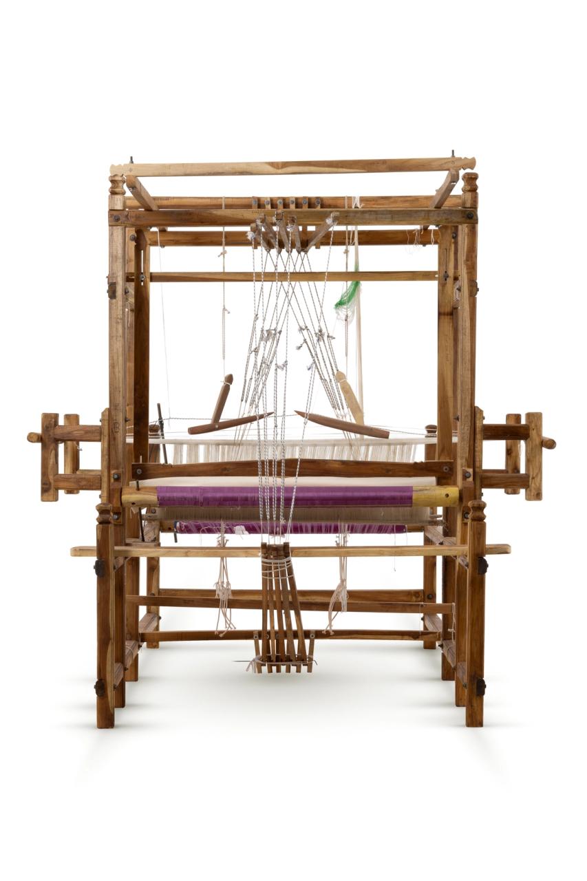 A sturdy wooden frame loom with five pedals. The loom has been warped with purple silk which is partially woven in stripes with the same purple silk and gold thread. The cloth is patterned with a repeating diamond motif in gold on purple and purple on gold.