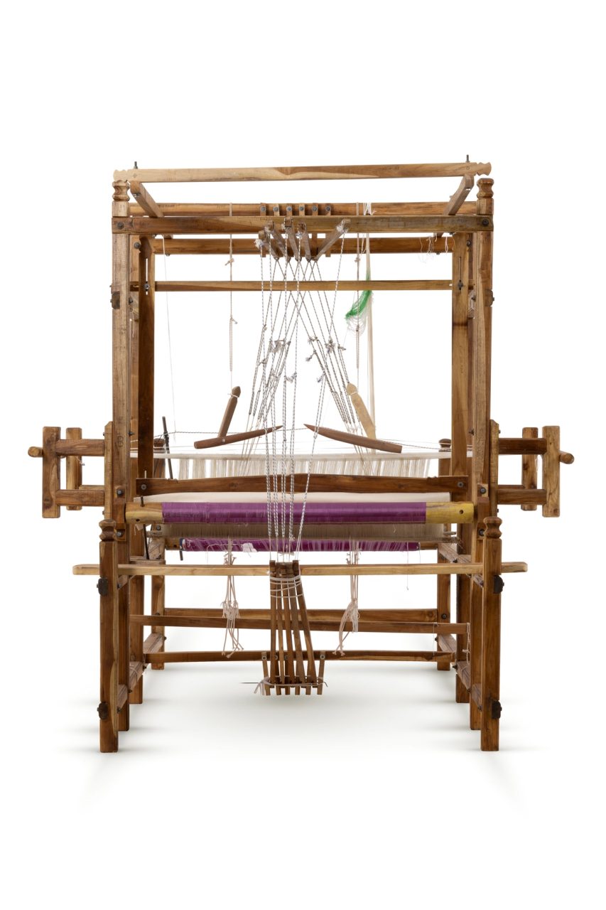 A sturdy wooden frame loom with five pedals. The loom has been warped with purple silk which is partially woven in stripes with the same purple silk and gold thread. The cloth is patterned with a repeating diamond motif in gold on purple and purple on gold.