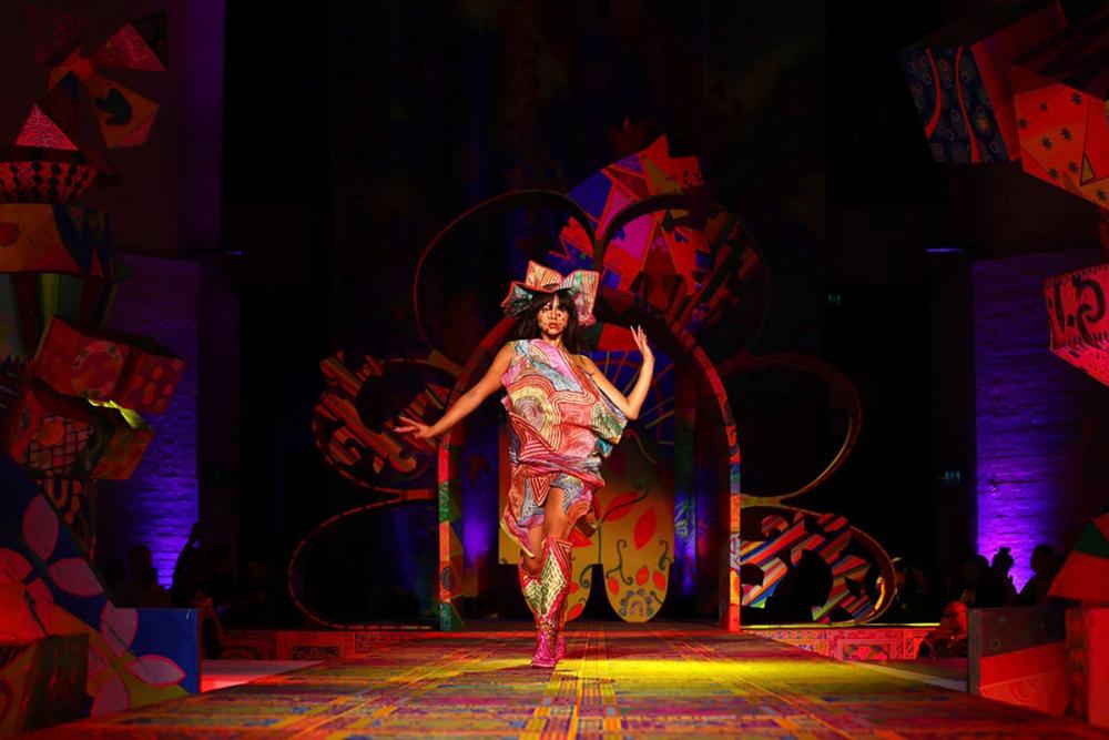 A model walks and poses on the runway wearing a multicoloured outfit and hat.