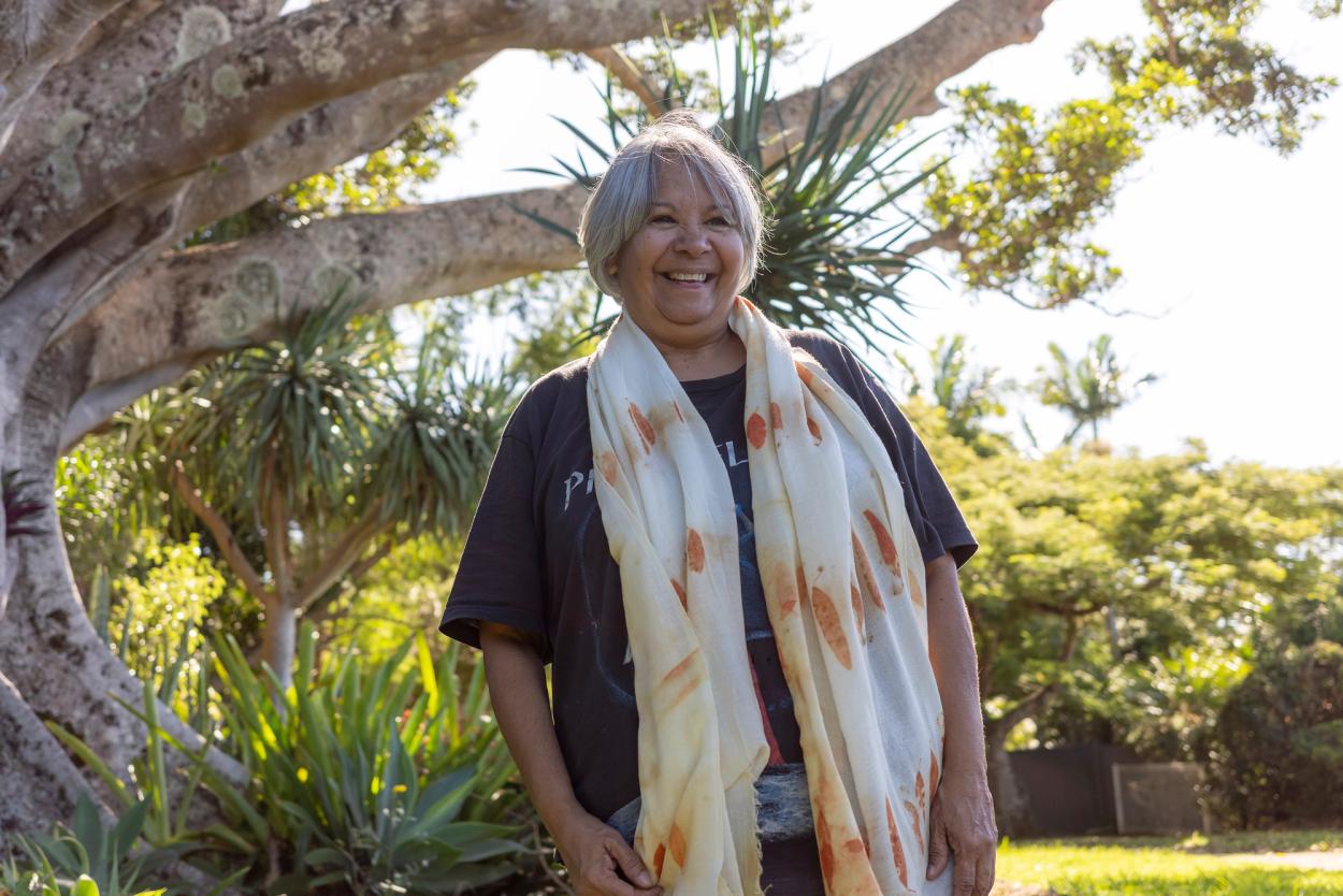 A smiling woman standing outside wearing a black t-shirt and a scarf with orange printed eucalyptus leaves draped around her neck.