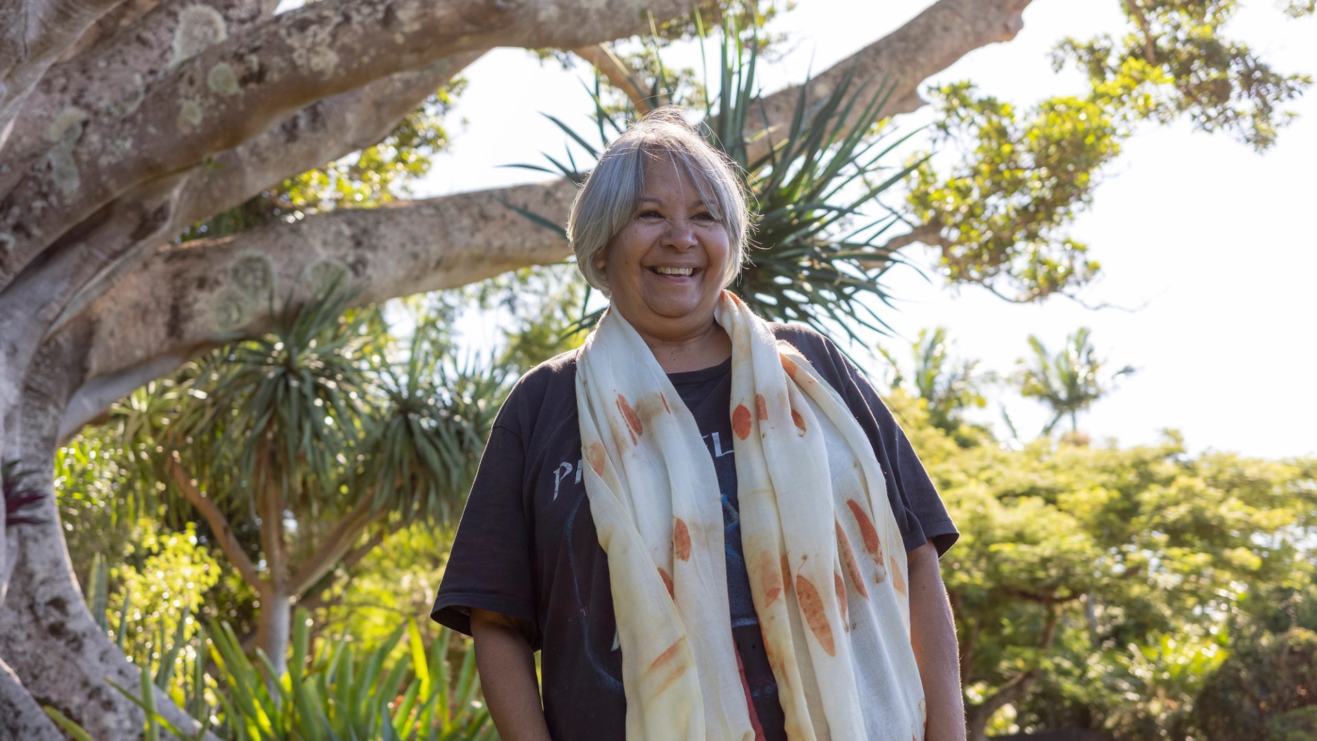 A smiling woman standing outside wearing a black t-shirt and a scarf with orange printed eucalyptus leaves draped around her neck.