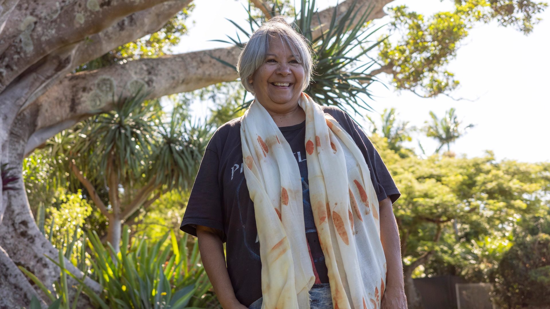 A smiling woman standing outside wearing a black t-shirt and a scarf with orange printed eucalyptus leaves draped around her neck.