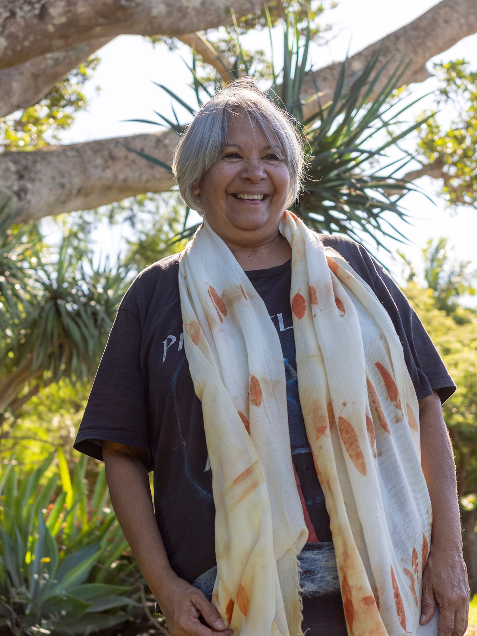 A smiling woman standing outside wearing a black t-shirt and a scarf with orange printed eucalyptus leaves draped around her neck.