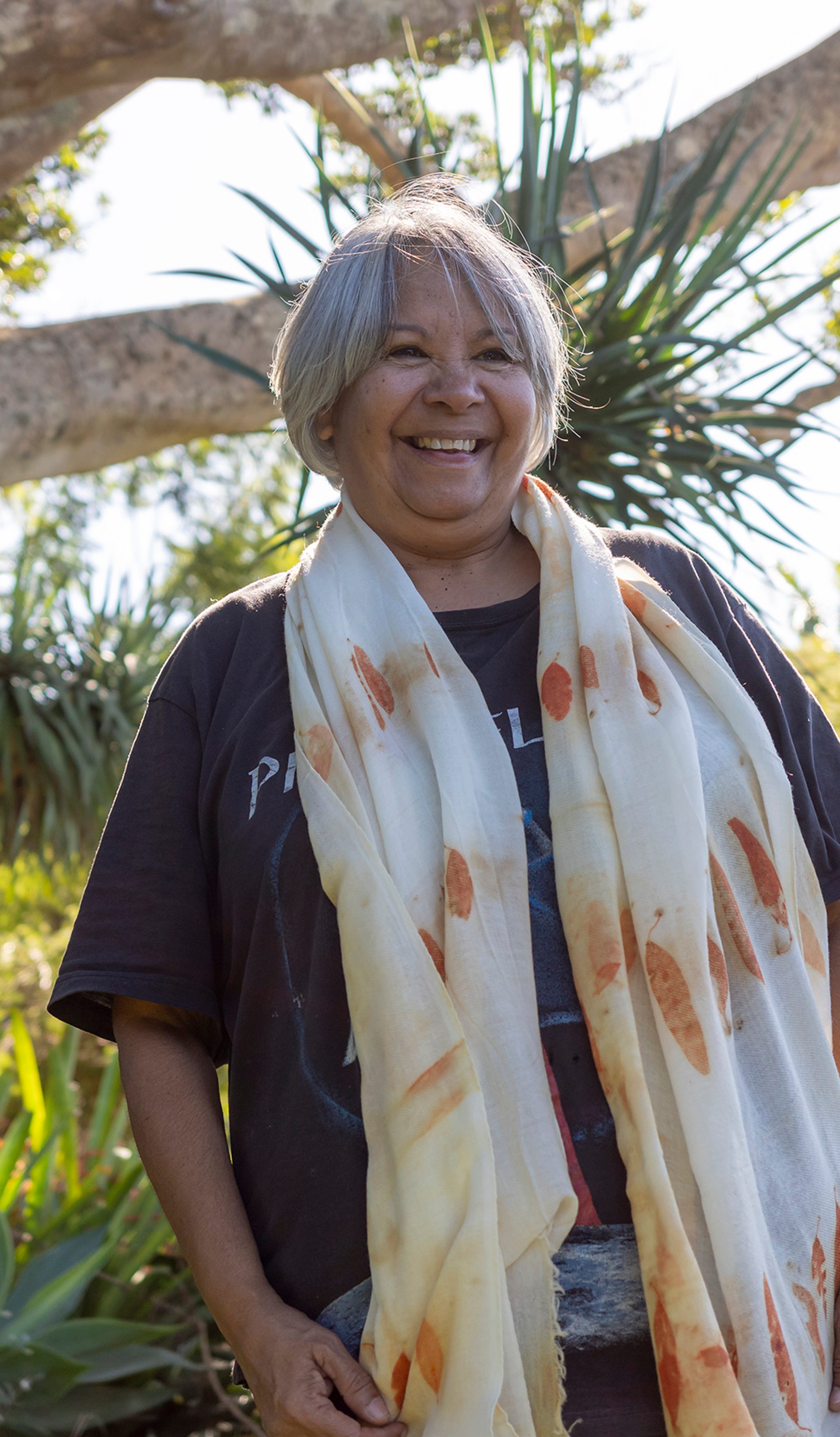 A smiling woman standing outside wearing a black t-shirt and a scarf with orange printed eucalyptus leaves draped around her neck.