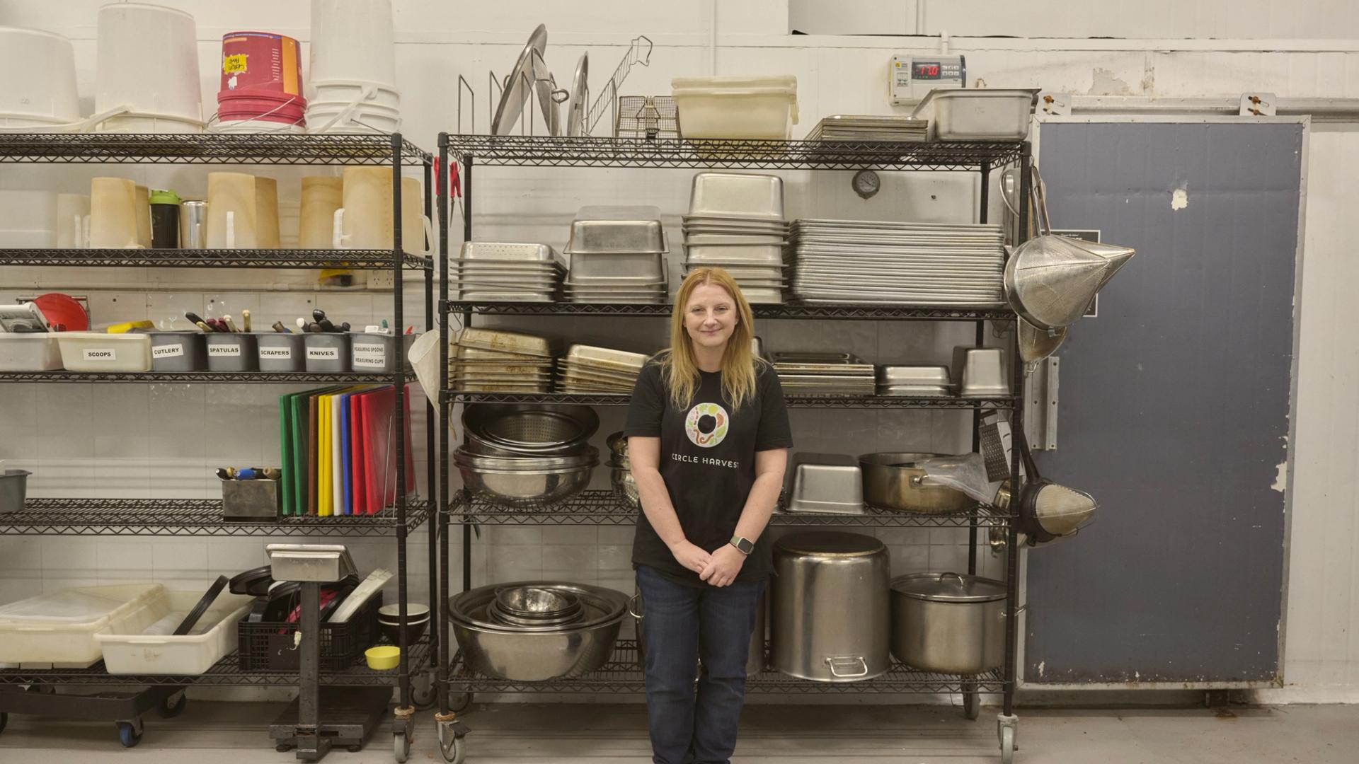 "A woman stands in front of a metal shelving unit filled with various kitchen equipment, including trays, pots, and utensils. She is wearing a black t-shirt with a colorful logo and jeans, smiling gently at the camera. The shelves are neatly organised, and the industrial kitchen setting has a clean, functional appearance."