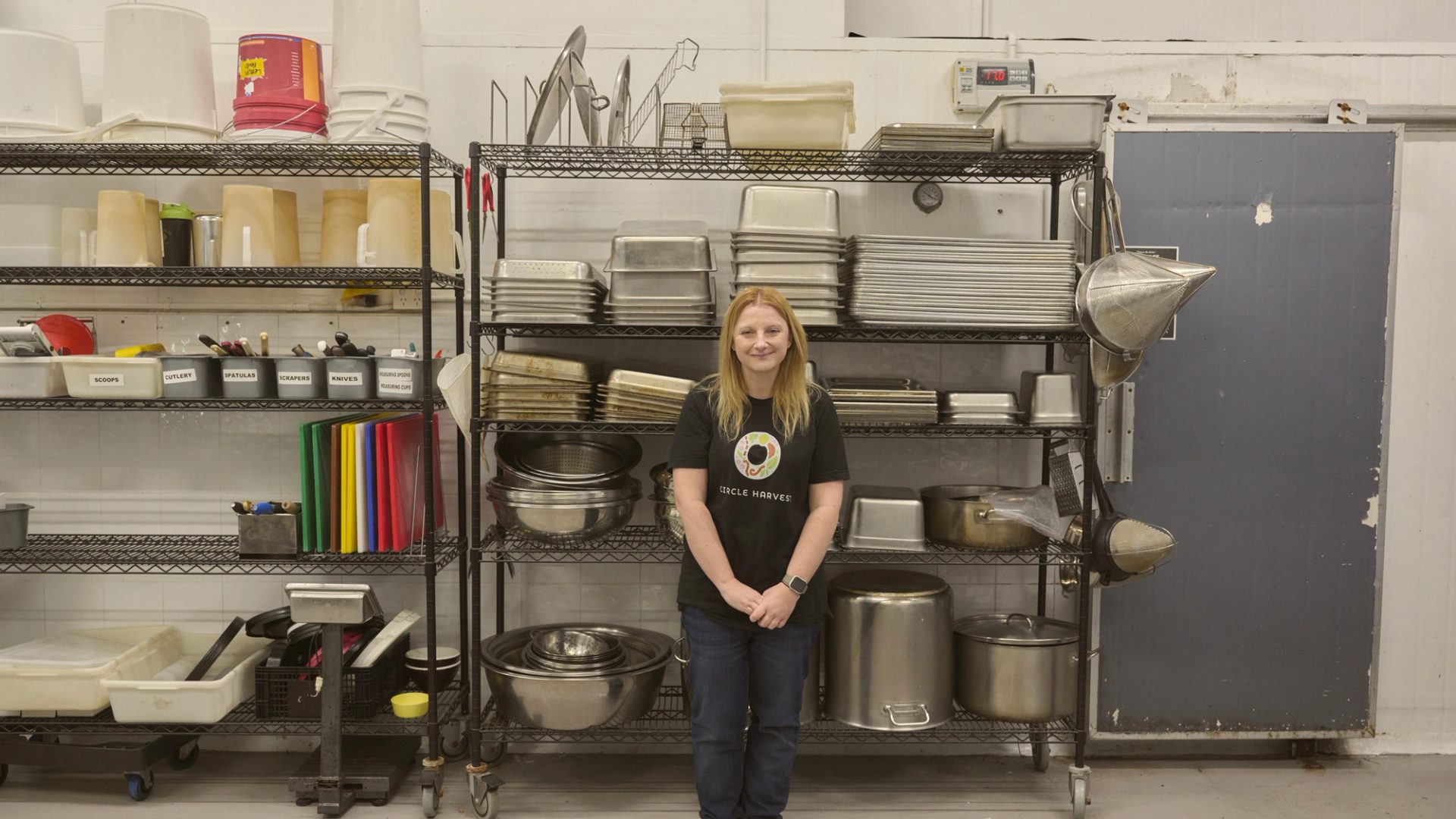 "A woman stands in front of a metal shelving unit filled with various kitchen equipment, including trays, pots, and utensils. She is wearing a black t-shirt with a colorful logo and jeans, smiling gently at the camera. The shelves are neatly organised, and the industrial kitchen setting has a clean, functional appearance."