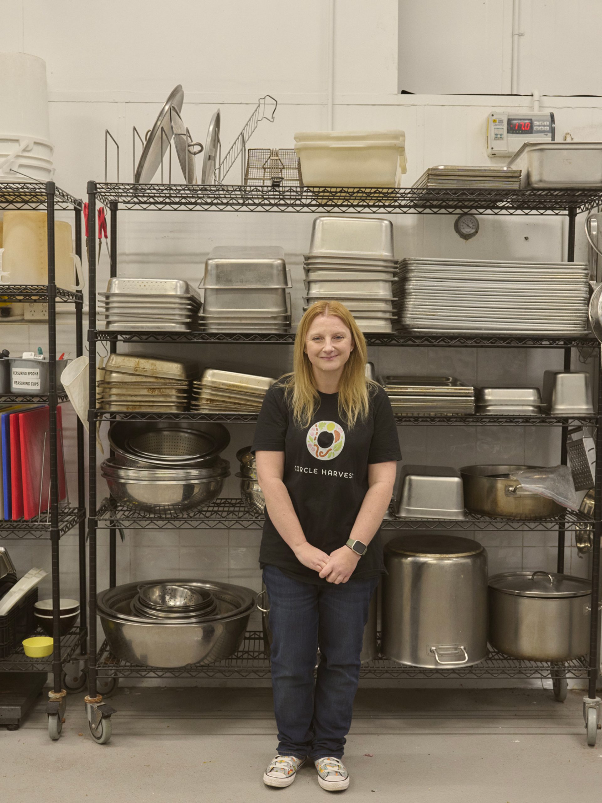 "A woman stands in front of a metal shelving unit filled with various kitchen equipment, including trays, pots, and utensils. She is wearing a black t-shirt with a colorful logo and jeans, smiling gently at the camera. The shelves are neatly organised, and the industrial kitchen setting has a clean, functional appearance."