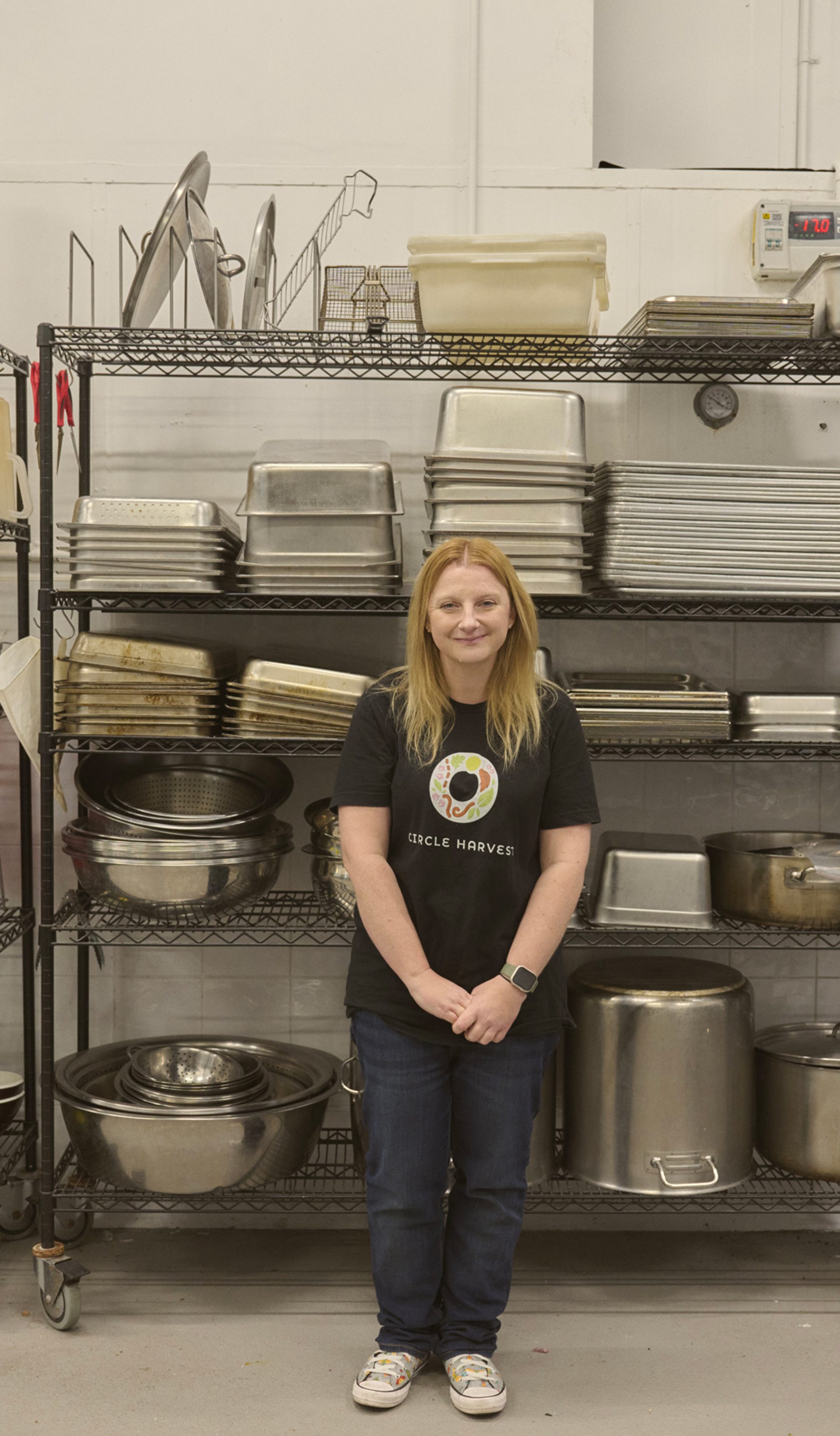 "A woman stands in front of a metal shelving unit filled with various kitchen equipment, including trays, pots, and utensils. She is wearing a black t-shirt with a colorful logo and jeans, smiling gently at the camera. The shelves are neatly organised, and the industrial kitchen setting has a clean, functional appearance."