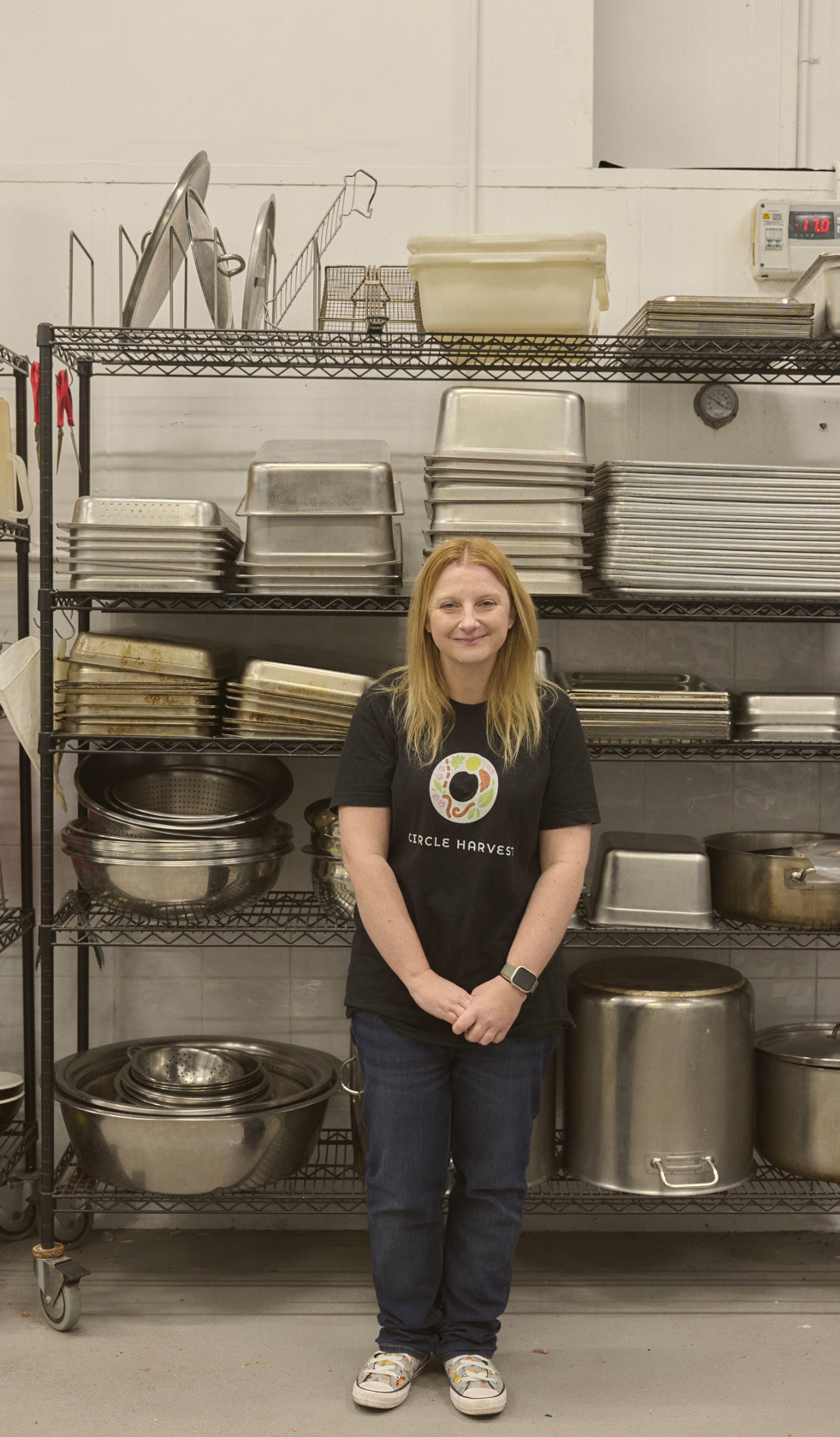 "A woman stands in front of a metal shelving unit filled with various kitchen equipment, including trays, pots, and utensils. She is wearing a black t-shirt with a colorful logo and jeans, smiling gently at the camera. The shelves are neatly organised, and the industrial kitchen setting has a clean, functional appearance."