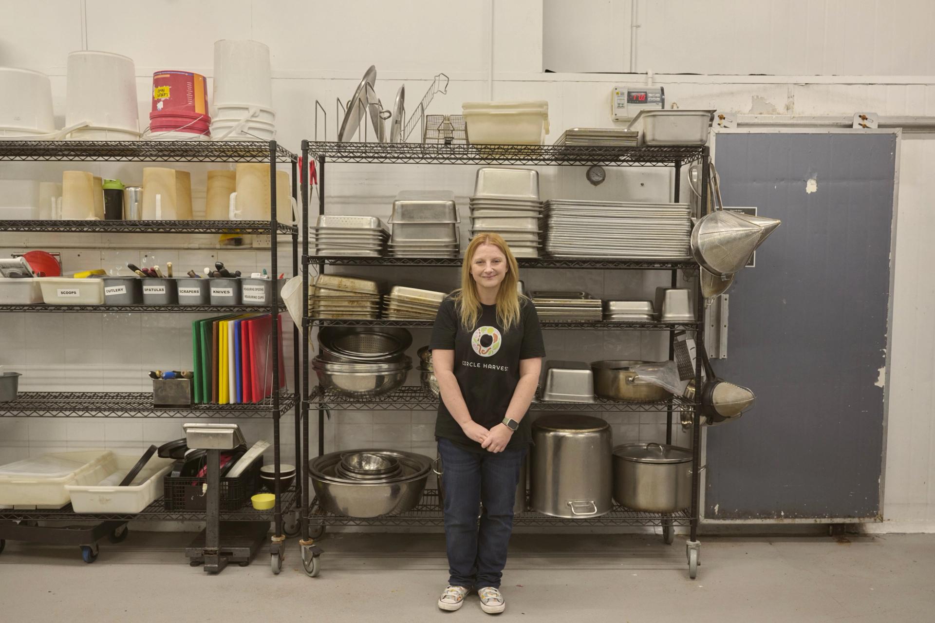 "A woman stands in front of a metal shelving unit filled with various kitchen equipment, including trays, pots, and utensils. She is wearing a black t-shirt with a colorful logo and jeans, smiling gently at the camera. The shelves are neatly organised, and the industrial kitchen setting has a clean, functional appearance."