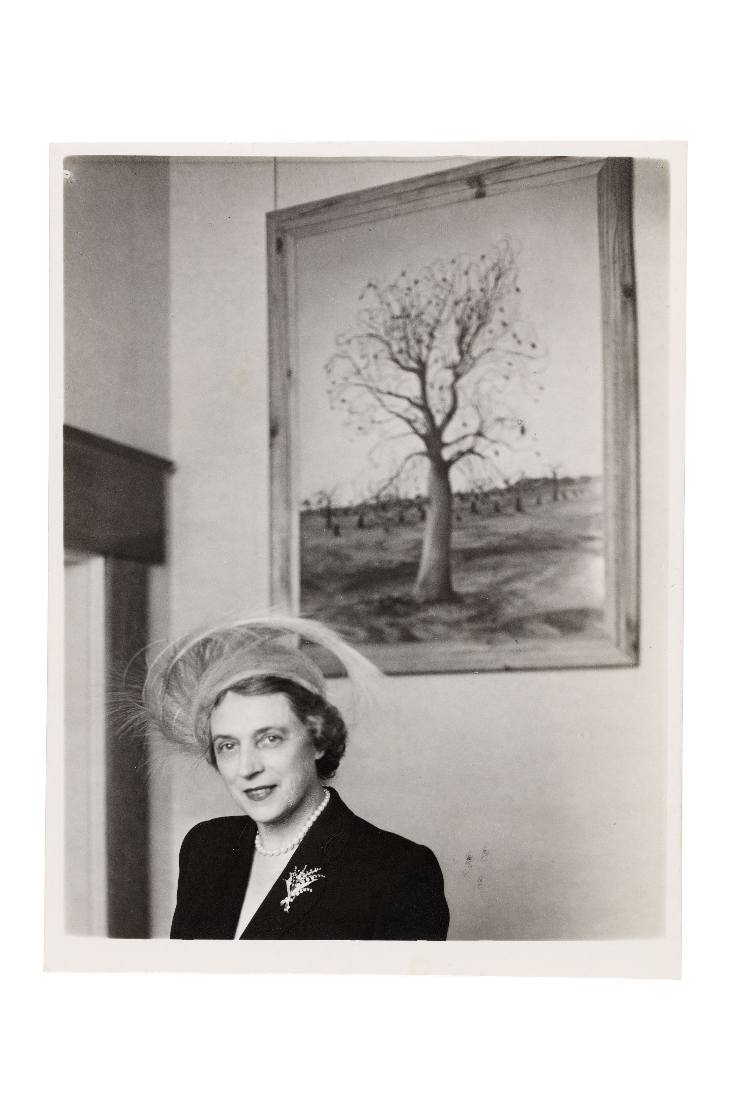 A woman, expensively dressed in the fashion of the time, stands in front of a painting of a tree.