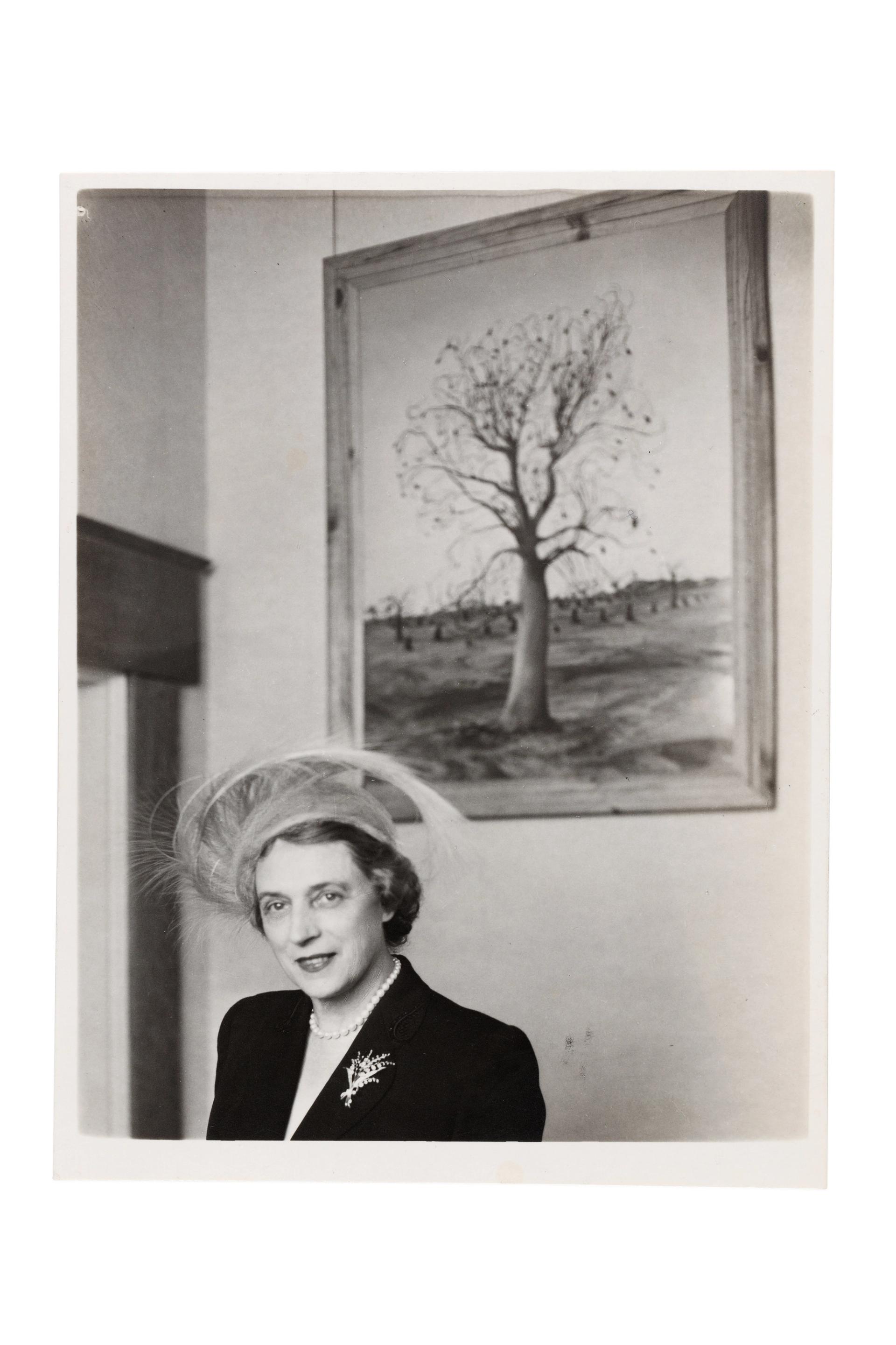 A woman, expensively dressed in the fashion of the time, stands in front of a painting of a tree.