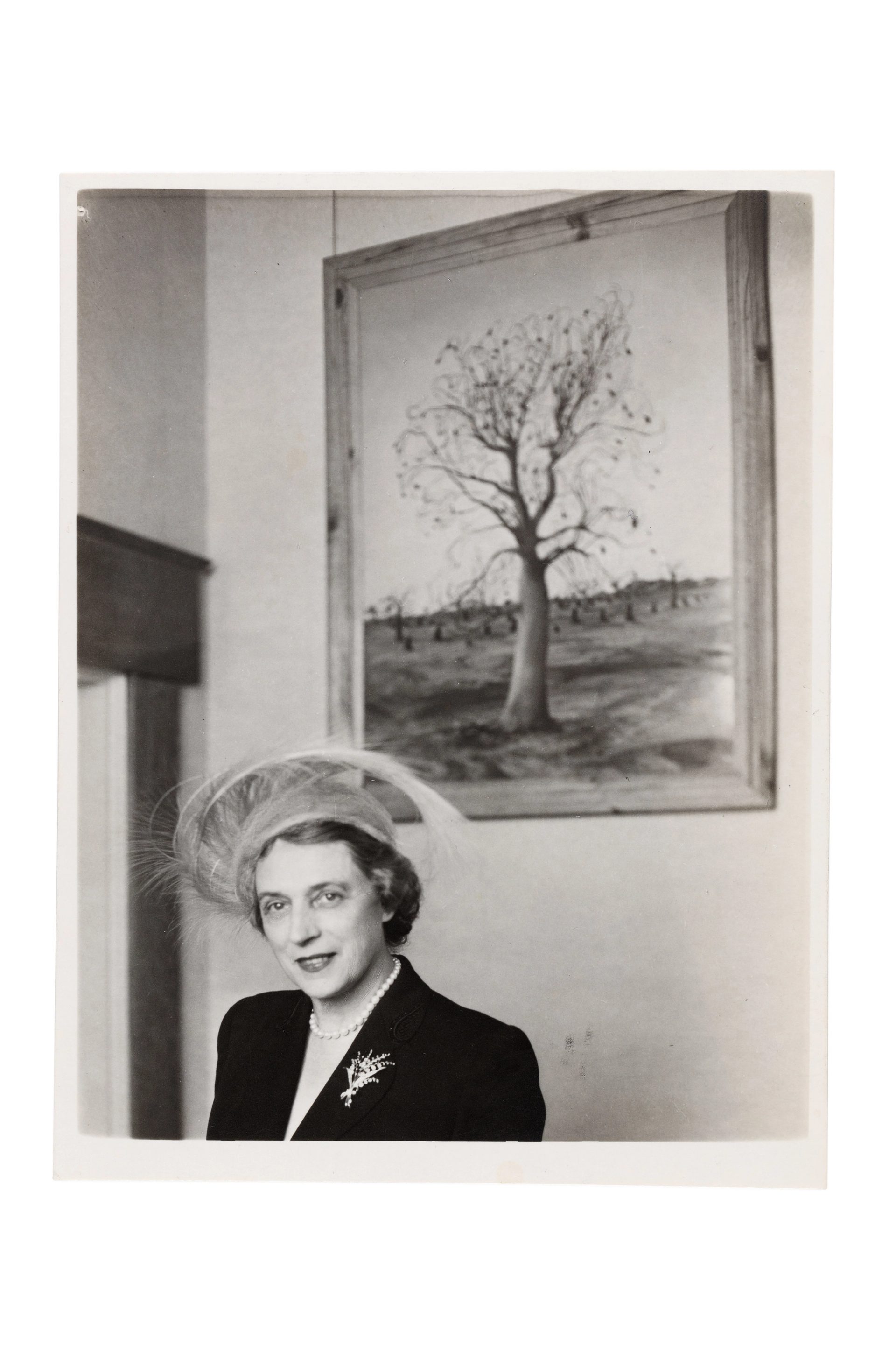 A woman, expensively dressed in the fashion of the time, stands in front of a painting of a tree.