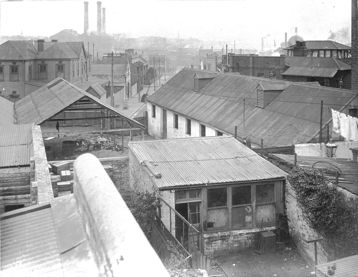 Black and white photograph of slums taken from a high vantage point with chimney stacks in the distance