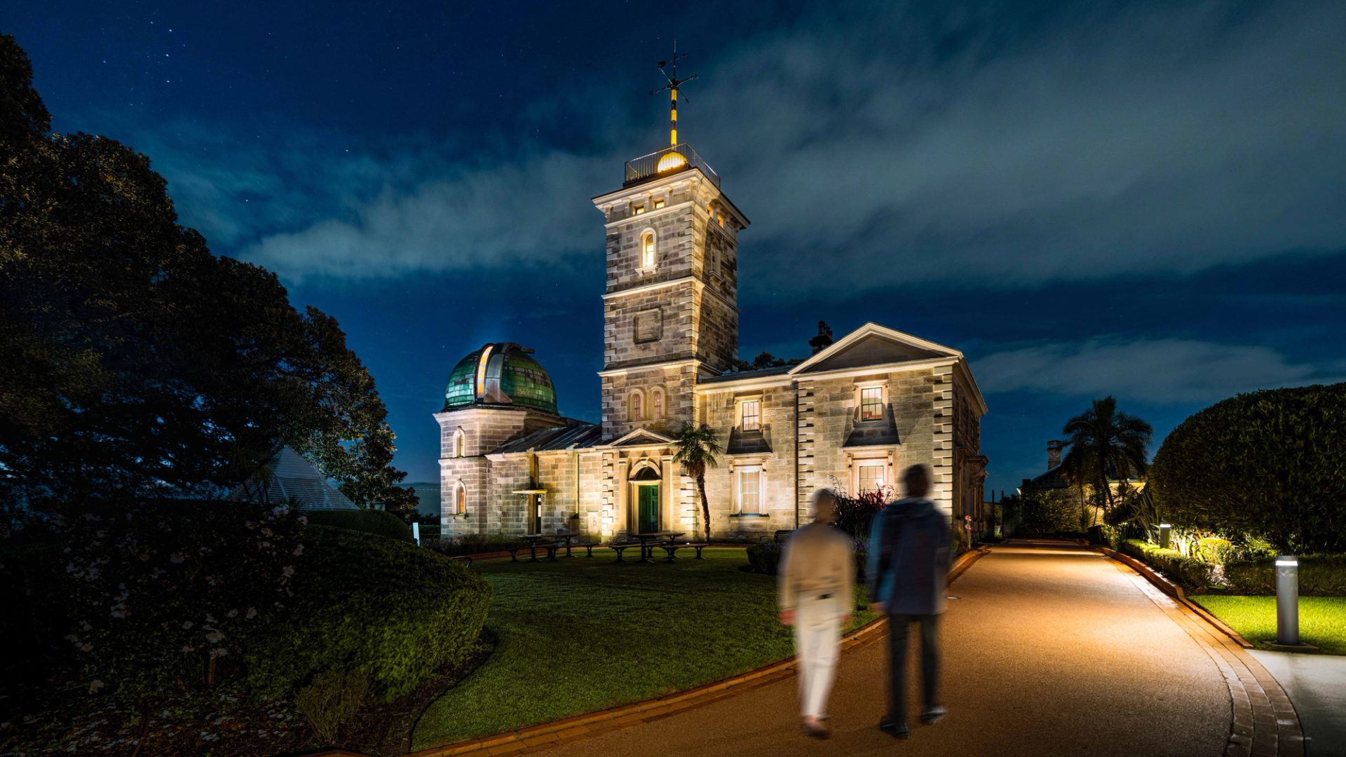 Two people walking down a driveway towards a heritage building.