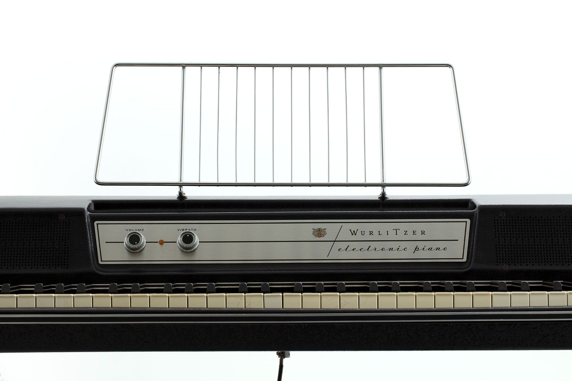 A cropped photo of the front of a black and silver keyboard. The front panel reads ‘Wurlitzer electric piano’. It has two dials and a metal music stand fixed on top.