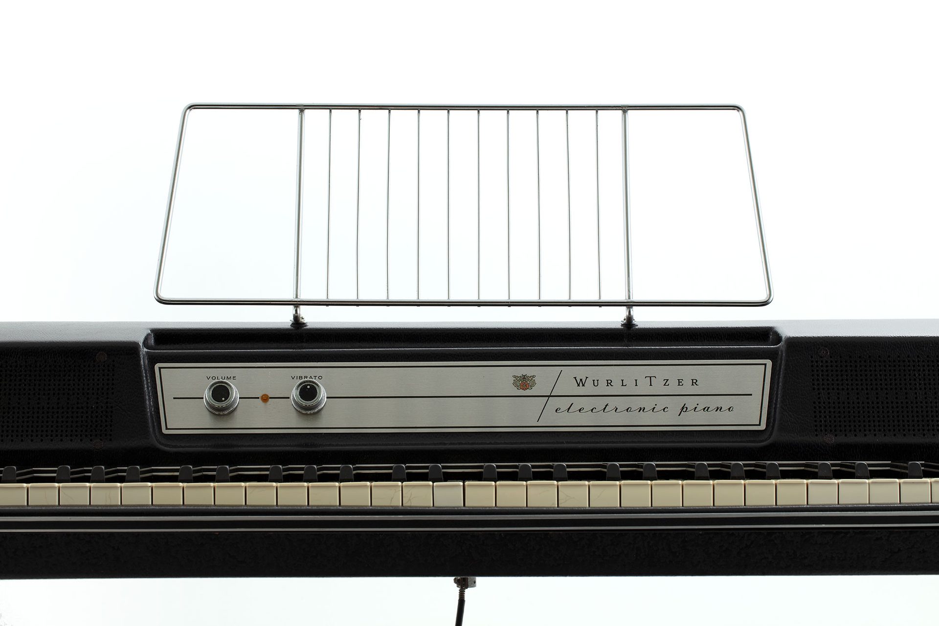 A cropped photo of the front of a black and silver keyboard. The front panel reads ‘Wurlitzer electric piano’. It has two dials and a metal music stand fixed on top.