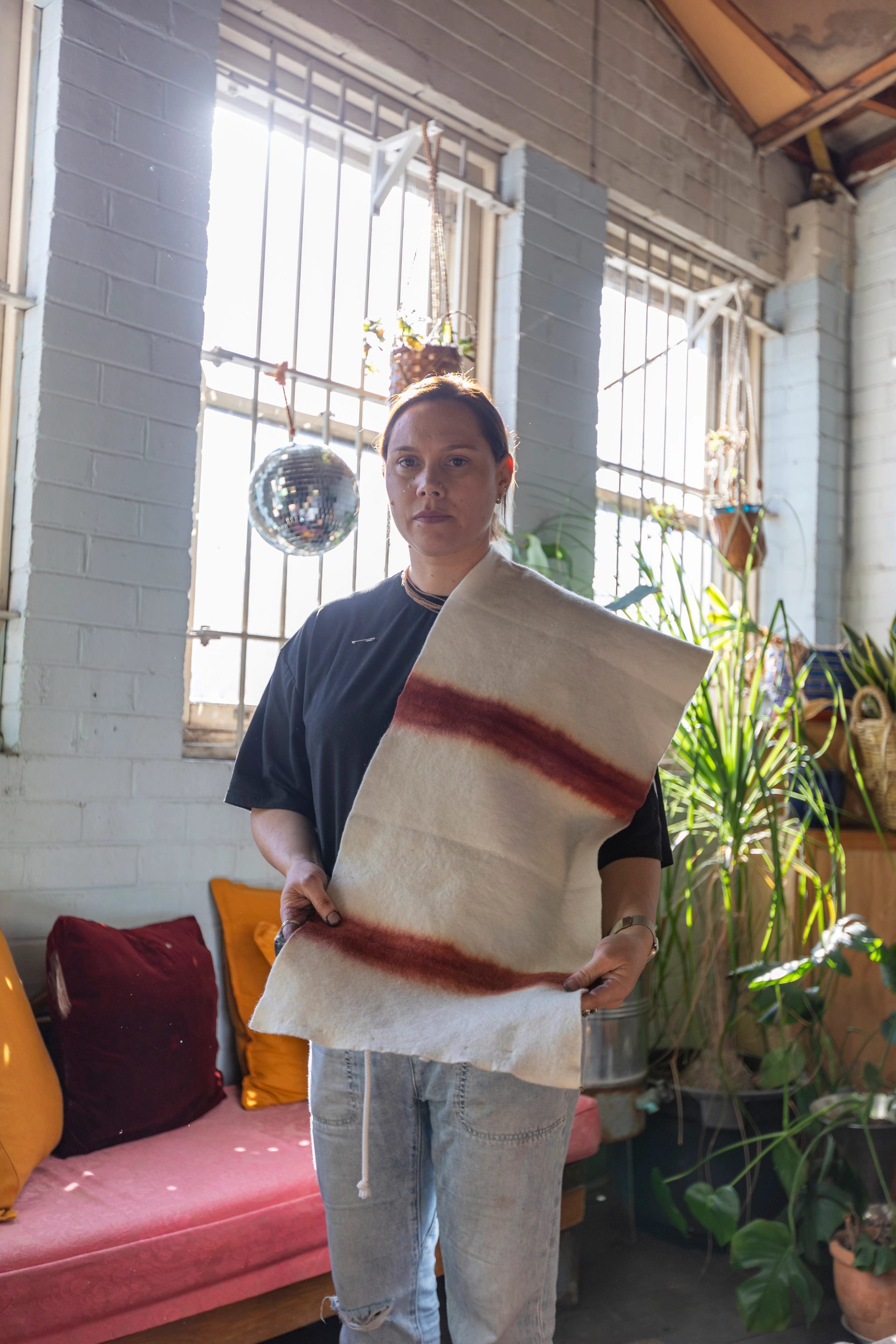 A woman standing in a room with light blue brick walls, a disco ball, plants and a red velvet couch. She is holding a piece of textile, with naturally-dyed red stripes.
