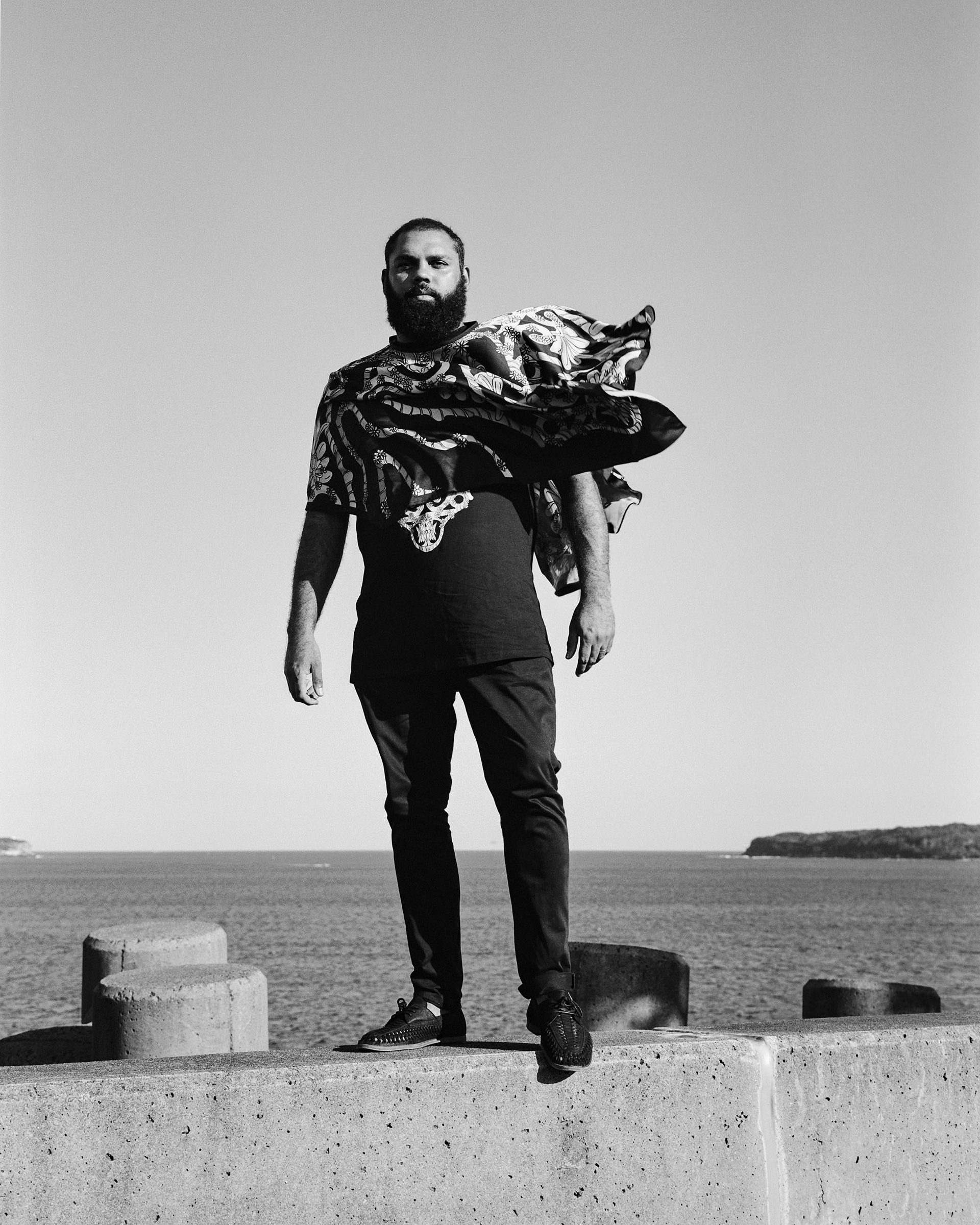 A man stands on a concrete barrier in front of Botany Bay. He is dressed in all black and wearing a large scarf across his shoulders that is blowing in the wind.