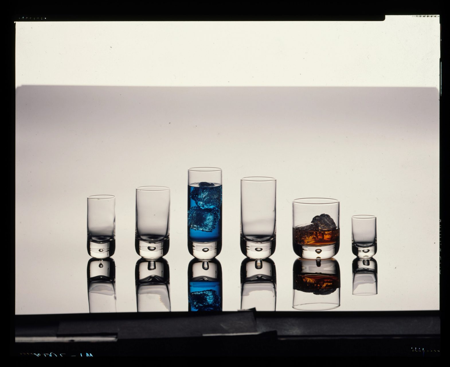 Photograph of six glasses of varying heights laid side by side on a reflective stand. The third and fifth glass from the left is filled with bright blue and amber liquid and ice cubes.