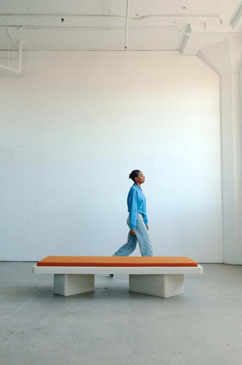 Orange cushion above a dynamic white daybed with figure wearing blue walking behind