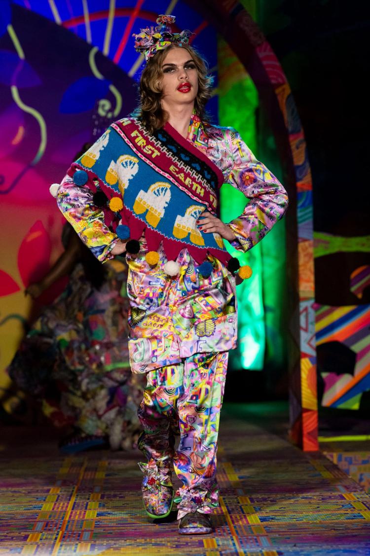 Runway model wearing colourful silk pyjama set with a large knitted sash with pom poms.