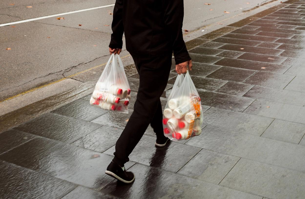 A figure wearing all black carries two plastic bags full of bottles.
