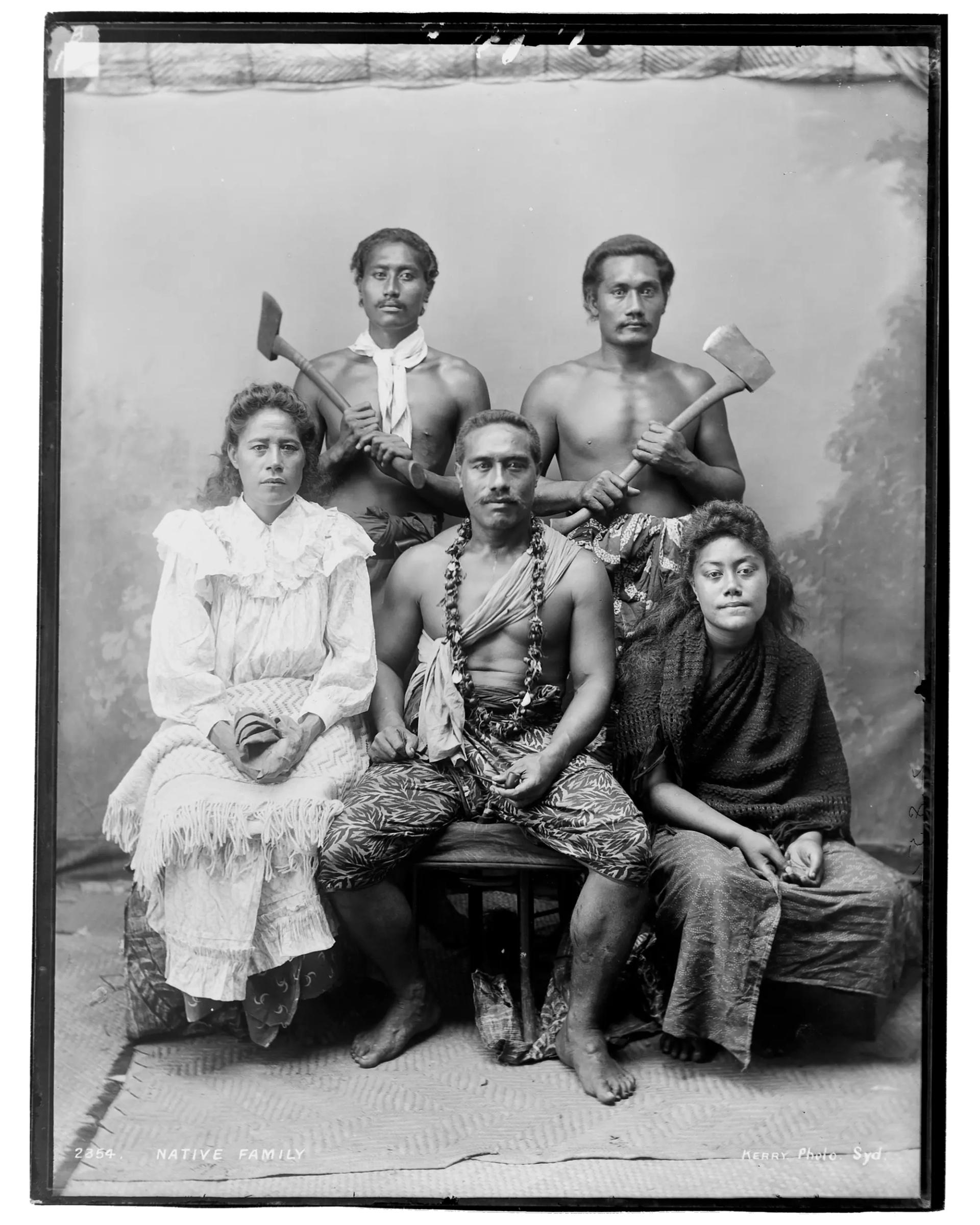 A silver gelatin dry plate glass negative in portrait format. The image shows a group of Pacific Islander people, three men and two women, photographed in a studio setting. The group has been photographed in front of a back drop painted with a forest scene. Two of the men stand holding axes. Seated on a bench in front of the standing men are two women and a man. The man sits in between the two women. The men wear wrap around fabric skirts. The seated man also wears a necklace and a scarf across his body. The woman in the left of the image wears a light coloured dress with a yoke and long, full sleeves. She holds a shawl in her lap. The woman in the right of the image wears a wrap around fabric skirt and a dark coloured shawl around her shoulders. Woven plant fibre mats cover the floor of the studio. The caption, studio number and studio mark are inscribed on the reverse of the negative.